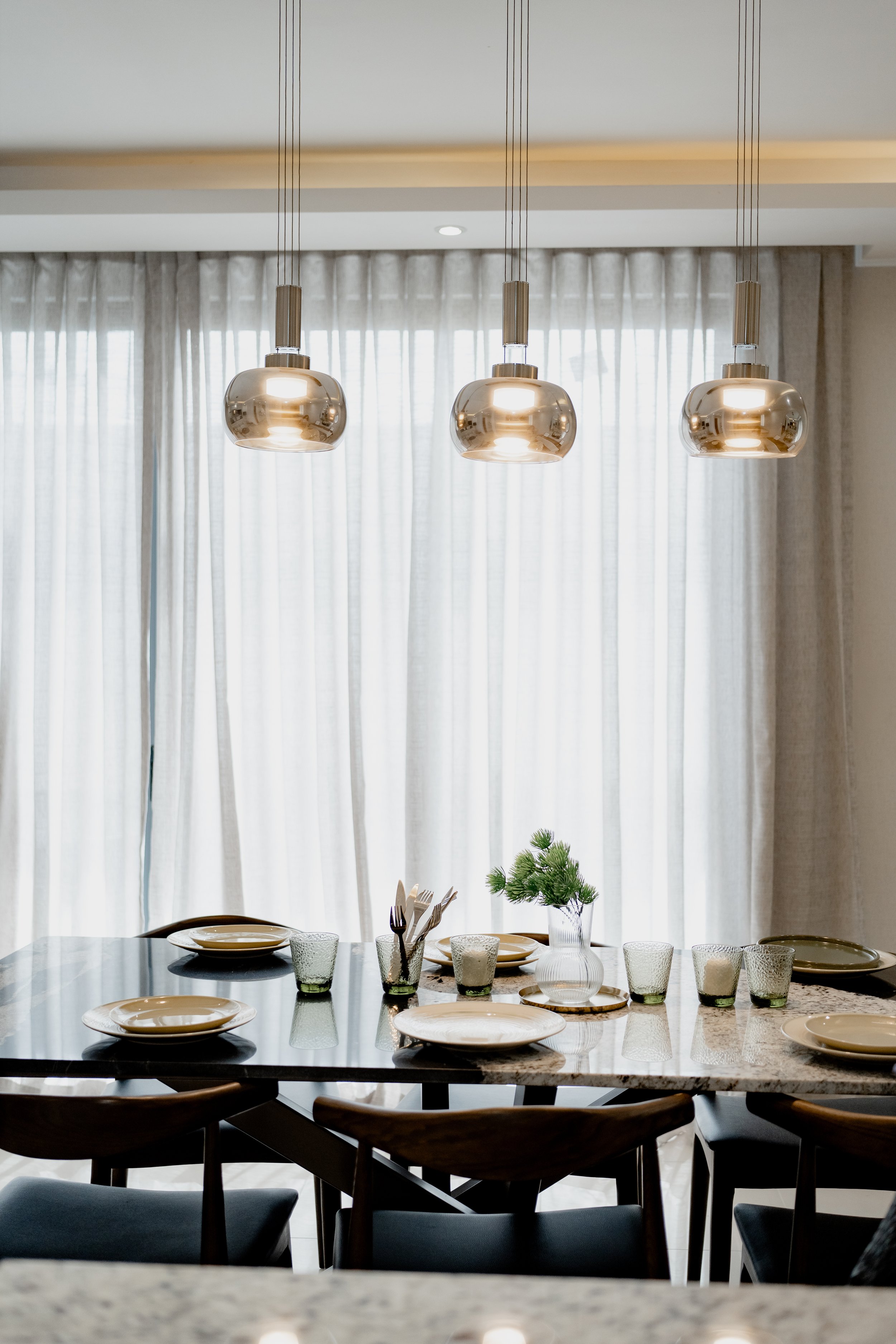 A modern dining room with a marble-top table set with cream-colored plates, glassware, and a small green potted plant, illuminated by three metallic pendant lights, with white curtains covering large windows in the background.