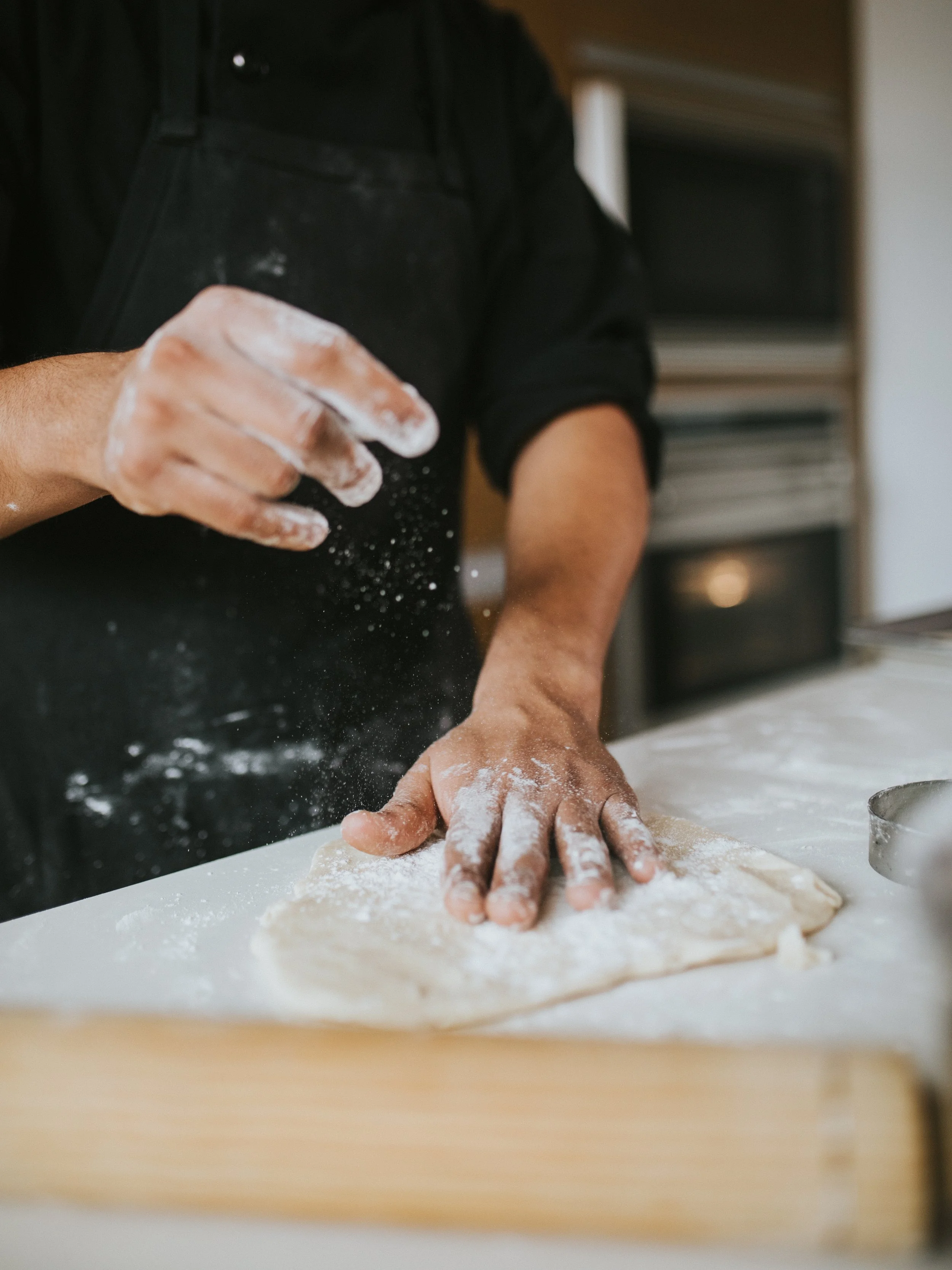 Person kneading dough on a floured surface with flour on their hands, wearing a black shirt, in a kitchen setting.