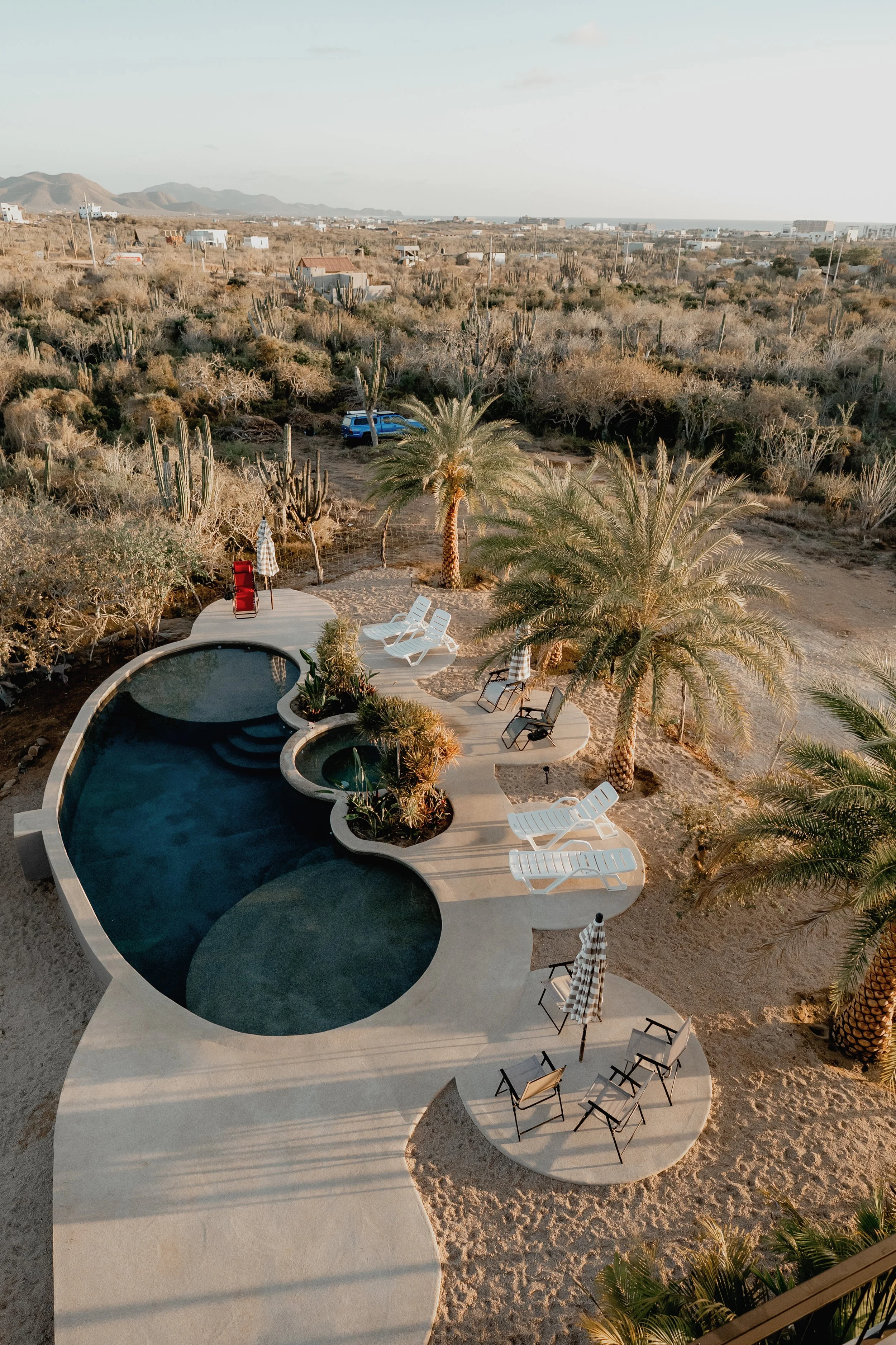 Aerial view of a backyard pool area with sun loungers, umbrellas, and palm trees, overlooking a desert landscape with cacti and distant mountains.