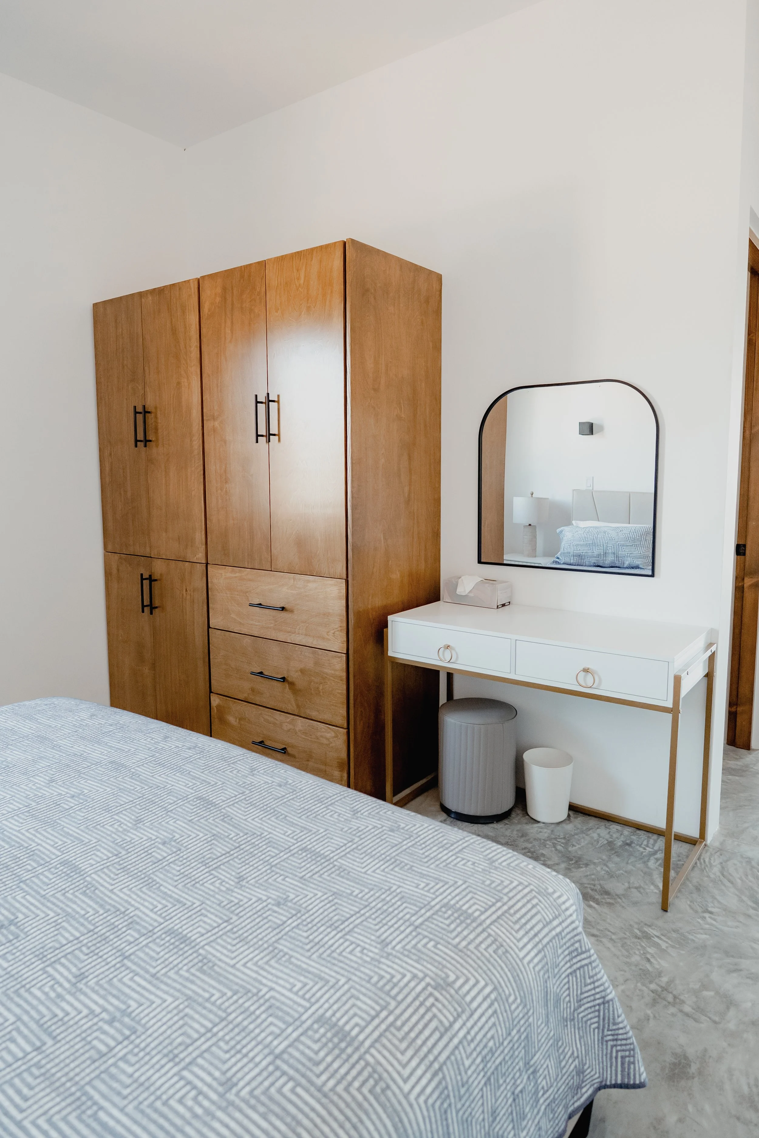 A bedroom with a wooden wardrobe, a white dresser with a round mirror, a bed with a patterned blue-and-white bedspread, a gray waste bin, a white paper waste basket, and a nightstand with a lamp and a wall-mounted light fixture.