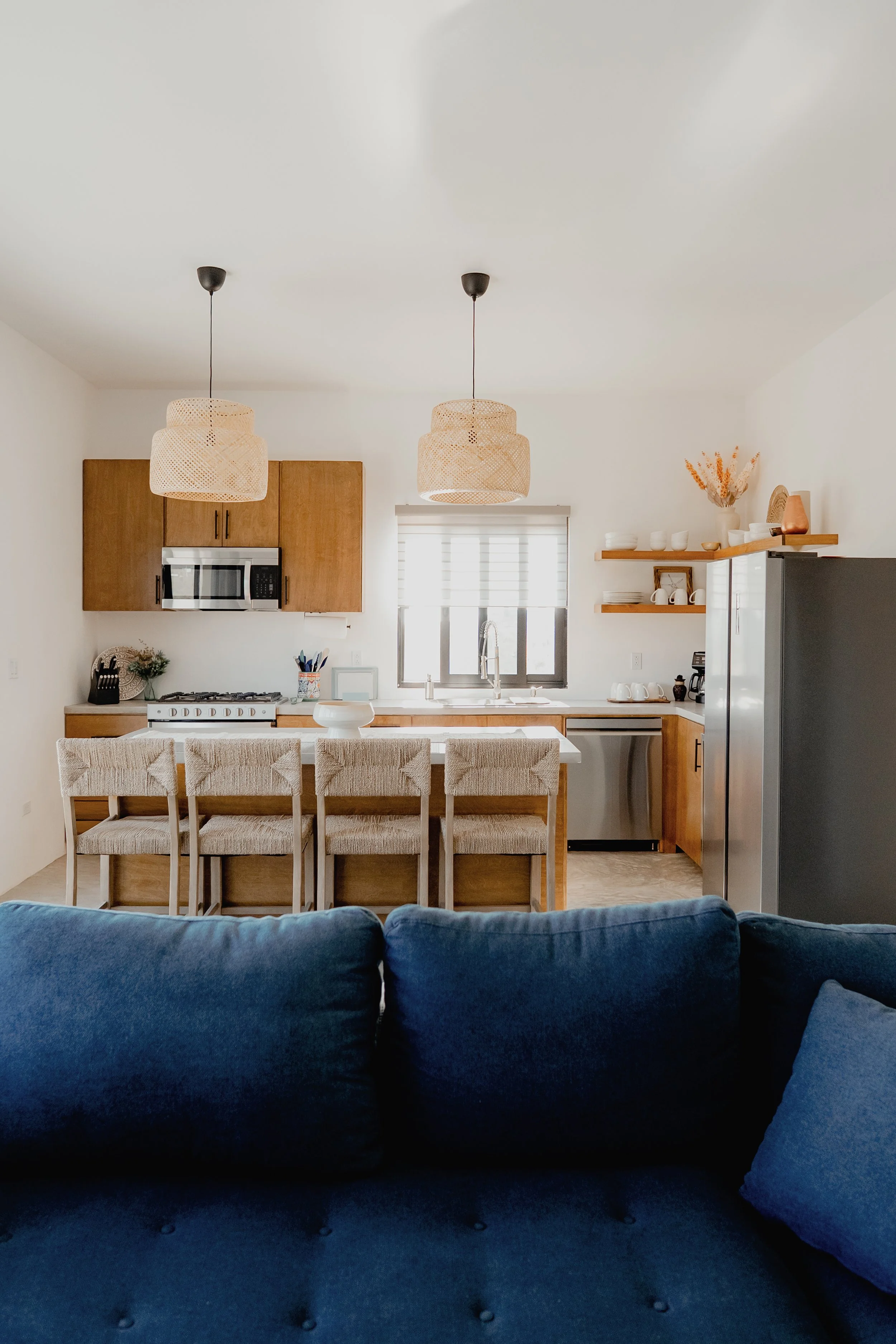 A modern kitchen and dining area with a navy blue sofa in the foreground. The kitchen has wooden cabinets, a white countertop, and a stainless steel fridge. Two woven pendant lights hang from the ceiling, and there are open wooden shelves with white 