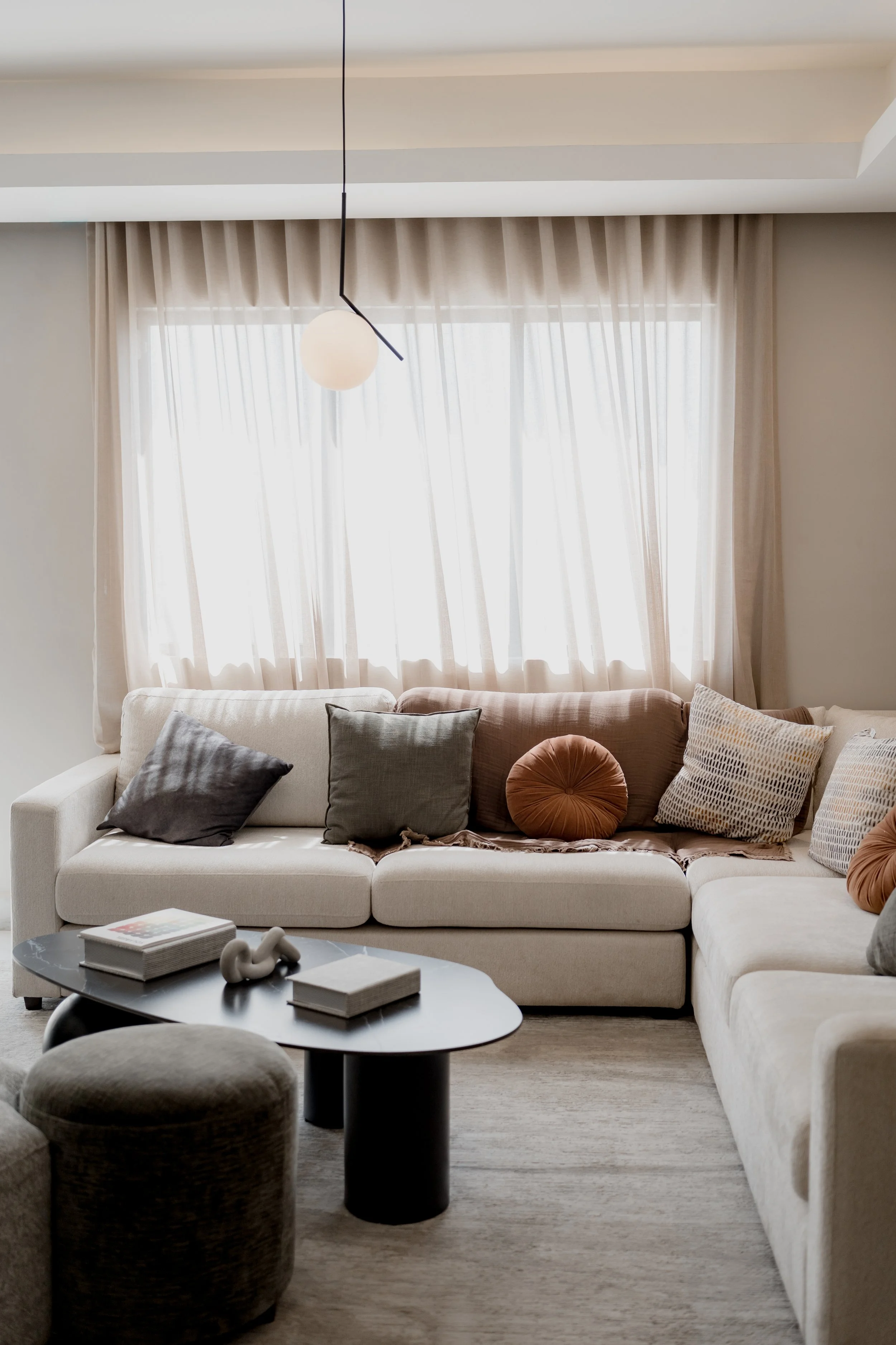 Living room with a beige sectional sofa, decorative pillows, and a black coffee table with books and ornaments, large windows with sheer curtains, and a modern hanging ceiling light.
