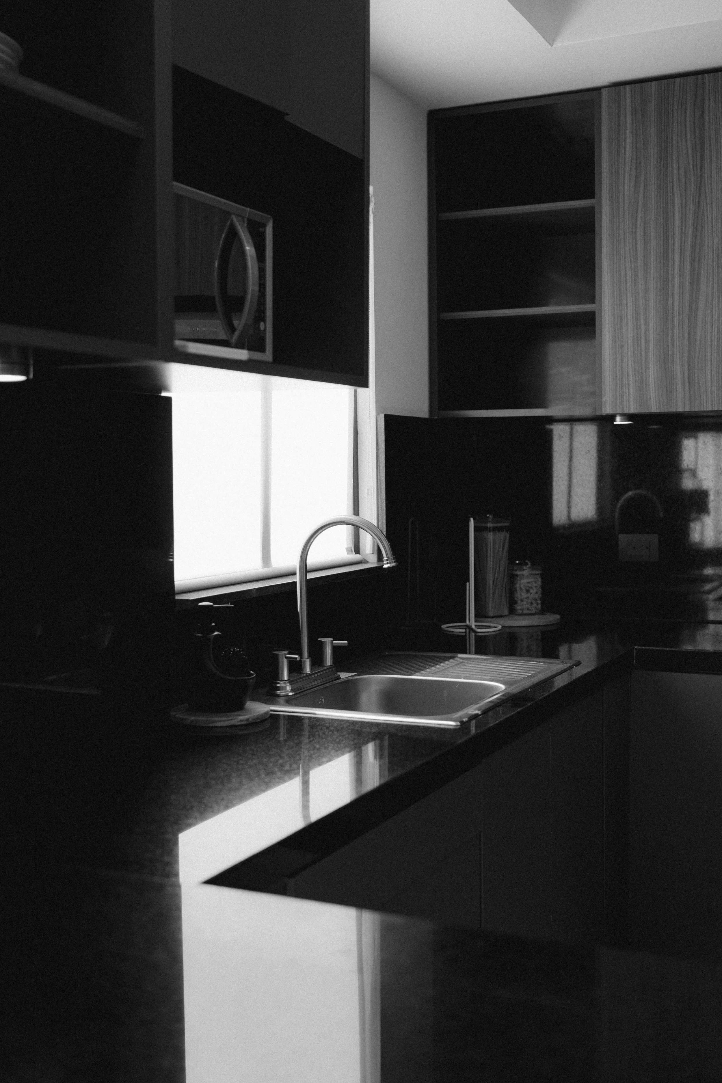A black and white photo of a modern kitchen with dark cabinets, a countertop, a sink with a faucet, a microwave oven, and some jars. There is a window above the countertop allowing natural light to illuminate the space.