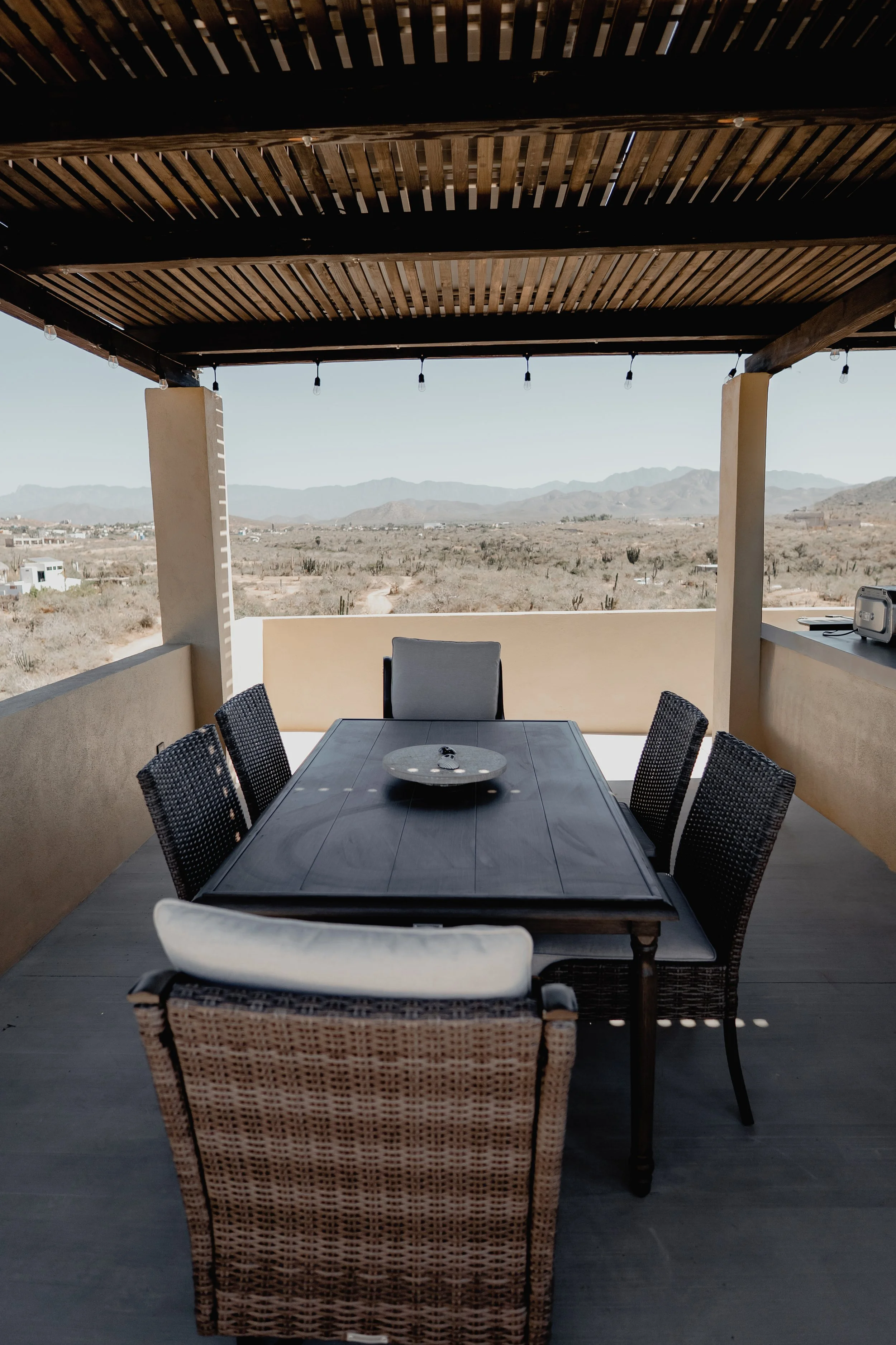 A covered outdoor patio with a dark wood slatted roof, overlooking a desert landscape with mountains in the background. The patio has a rectangular table with eight chairs around it and string lights hanging from the roof.