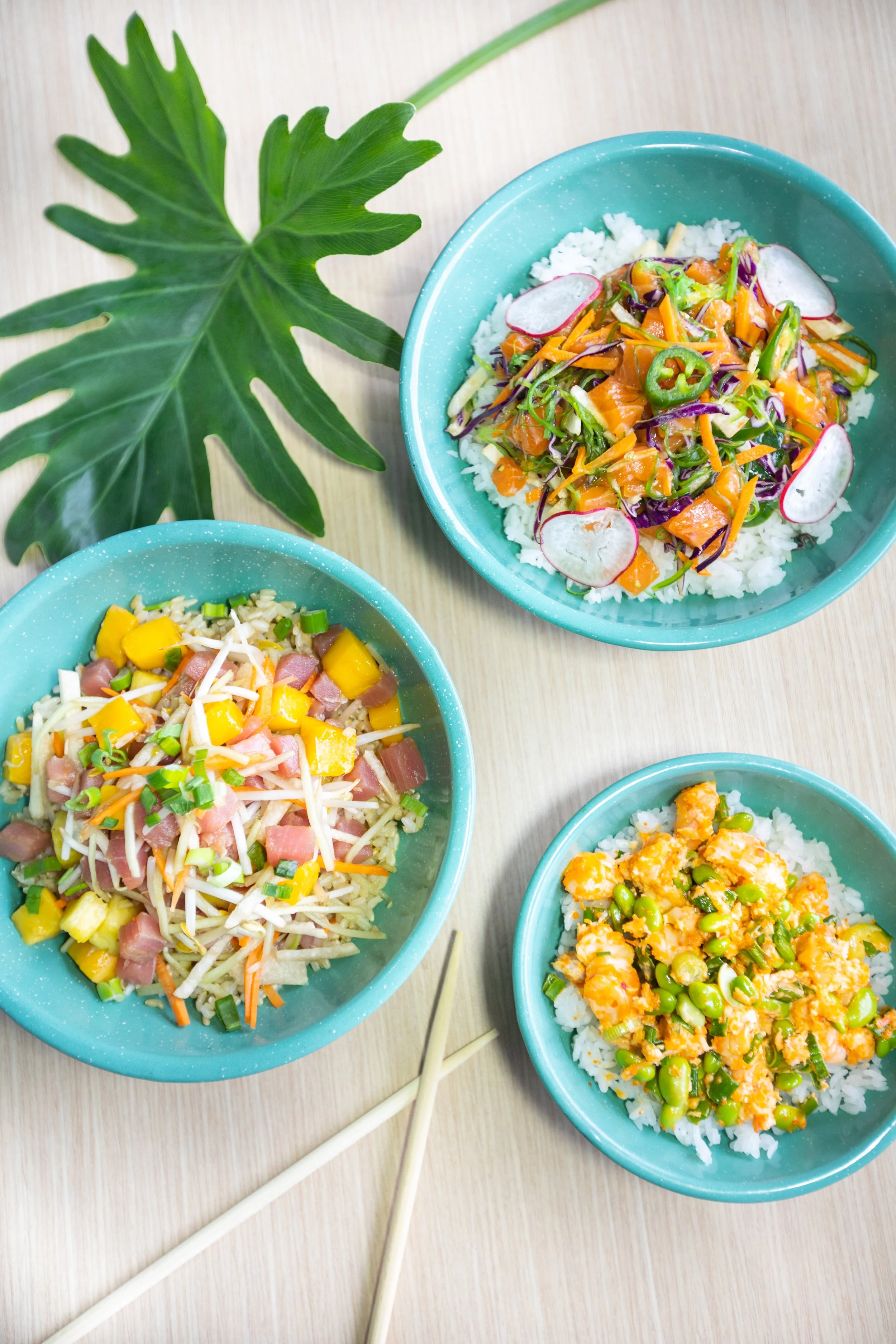 Three bowls of colorful Asian-inspired rice dishes with vegetables and seafood on a light wooden surface, accompanied by white chopsticks and a large green leaf.