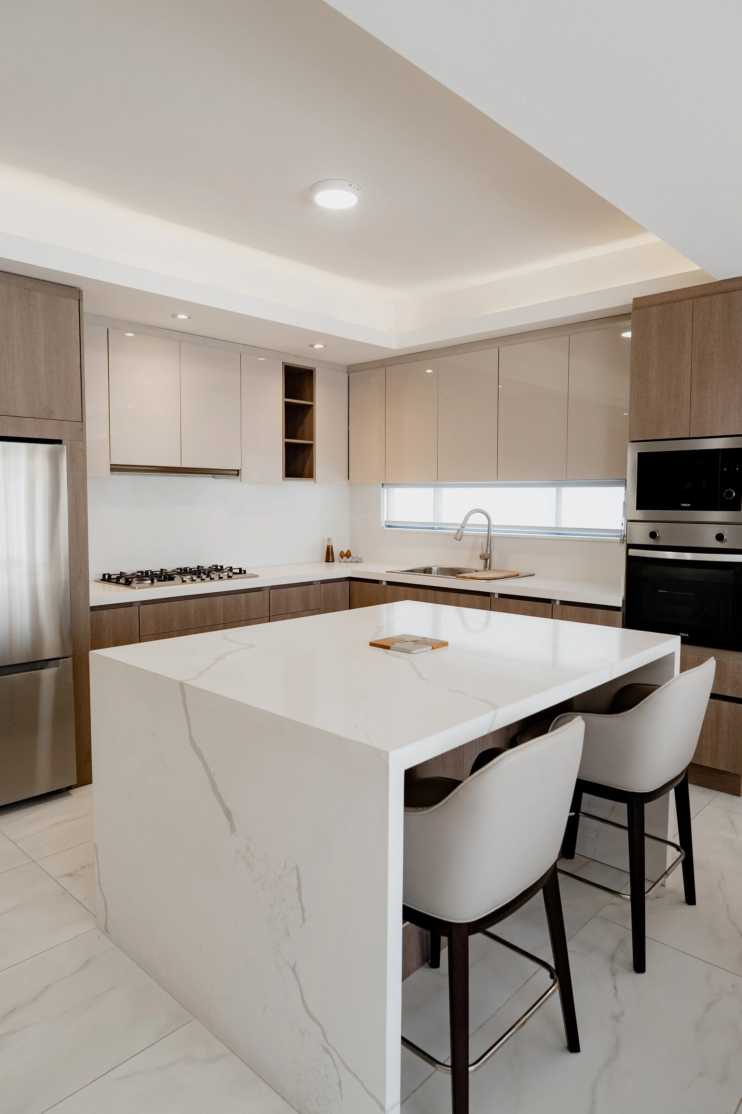 Modern kitchen with white marble island, beige upper cabinets, and stainless steel appliances, including a refrigerator and oven.
