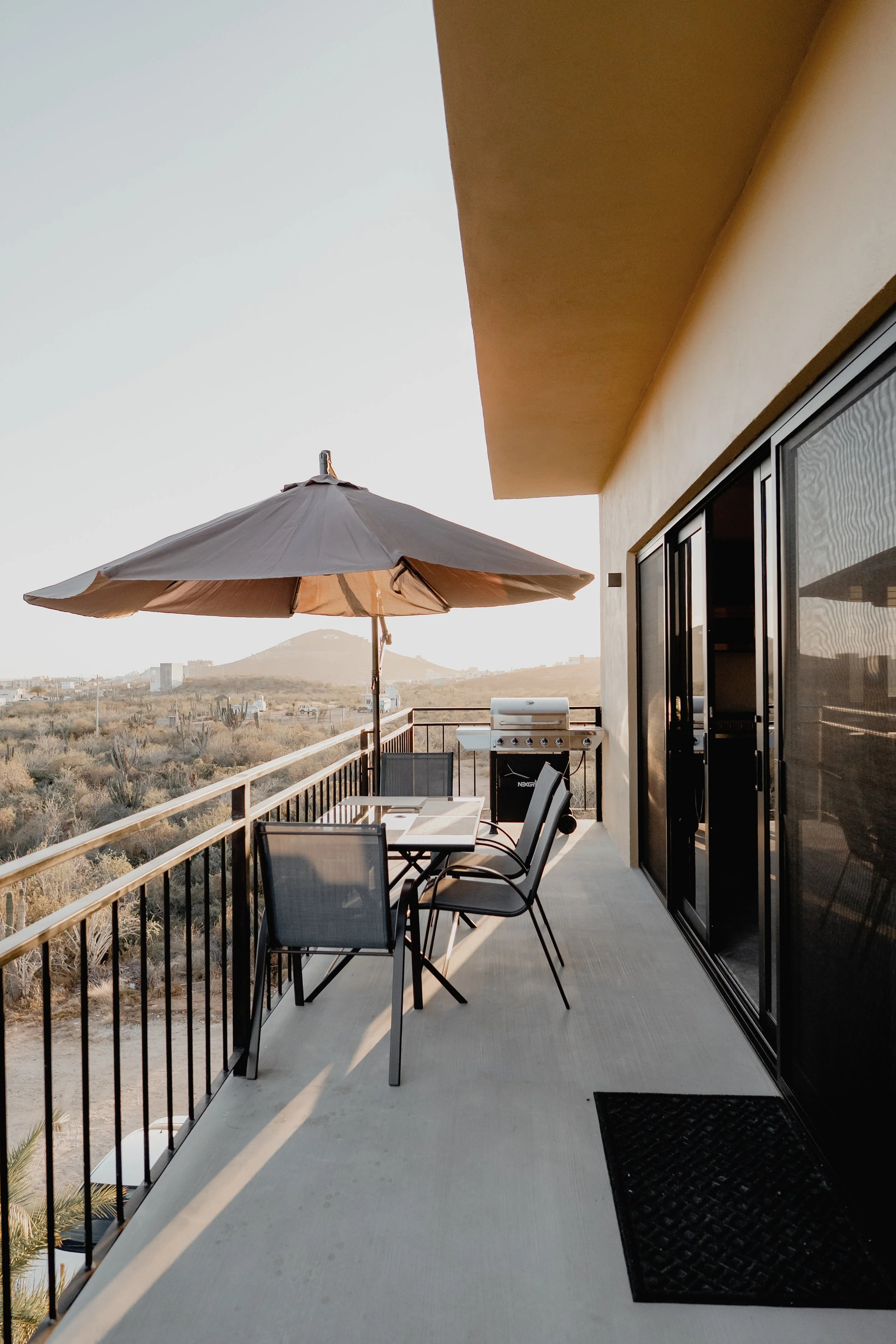 Balcony with outdoor furniture including a dining table, chairs, and an umbrella, overlooking a desert landscape with cacti and hills in the distance at sunset.