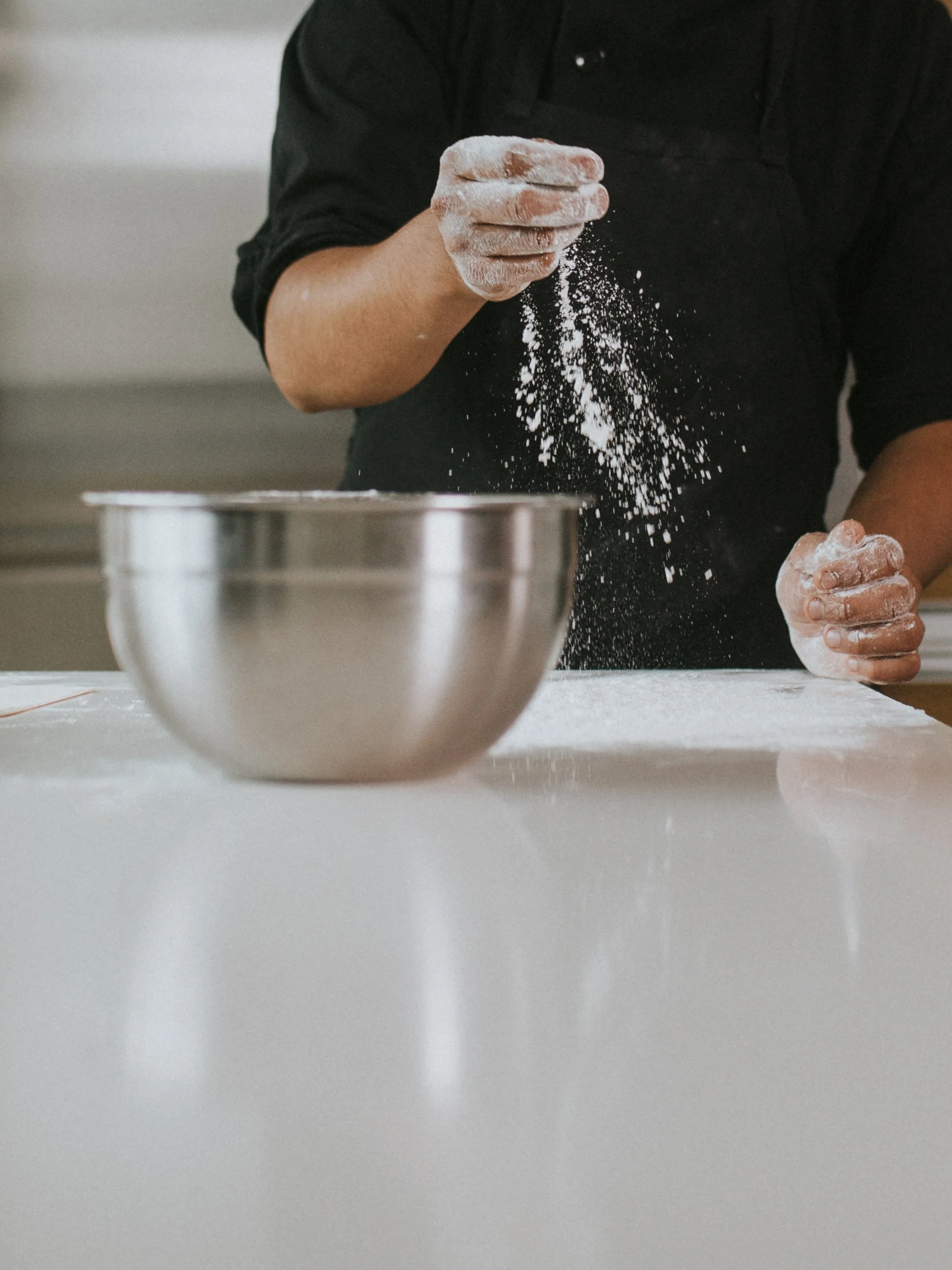 A person wearing a black shirt, with flour on their hands, is using their fingers to sift flour over a mixing bowl on a white kitchen counter.