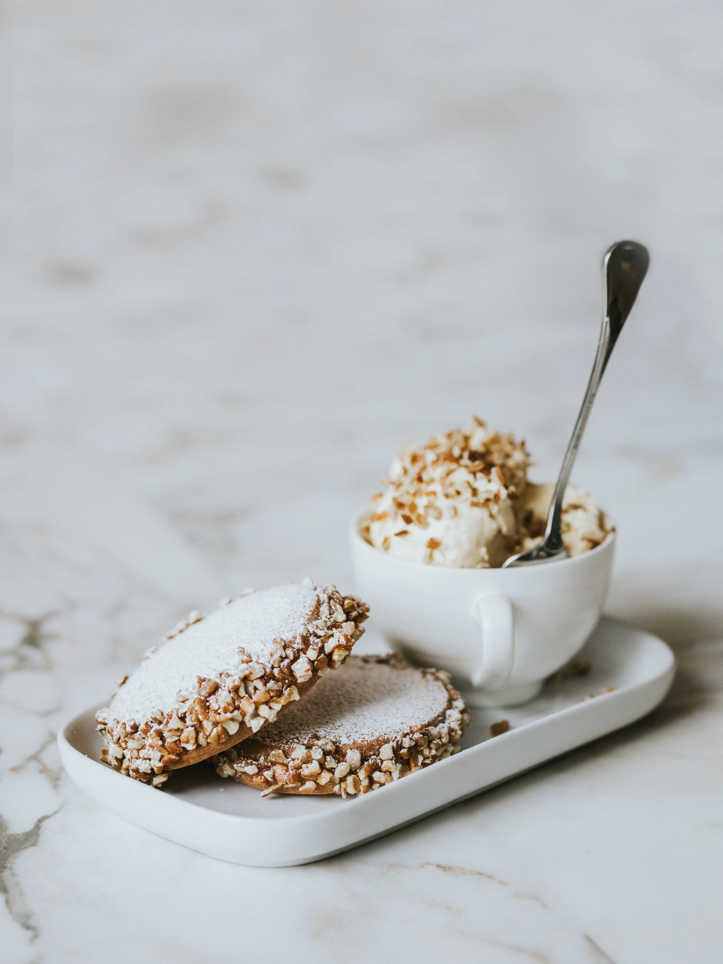 Close-up of a tray with chocolate cookies coated in chopped nuts and a cup of whipped cream topped with chocolate shavings and a spoon, on a marble surface.