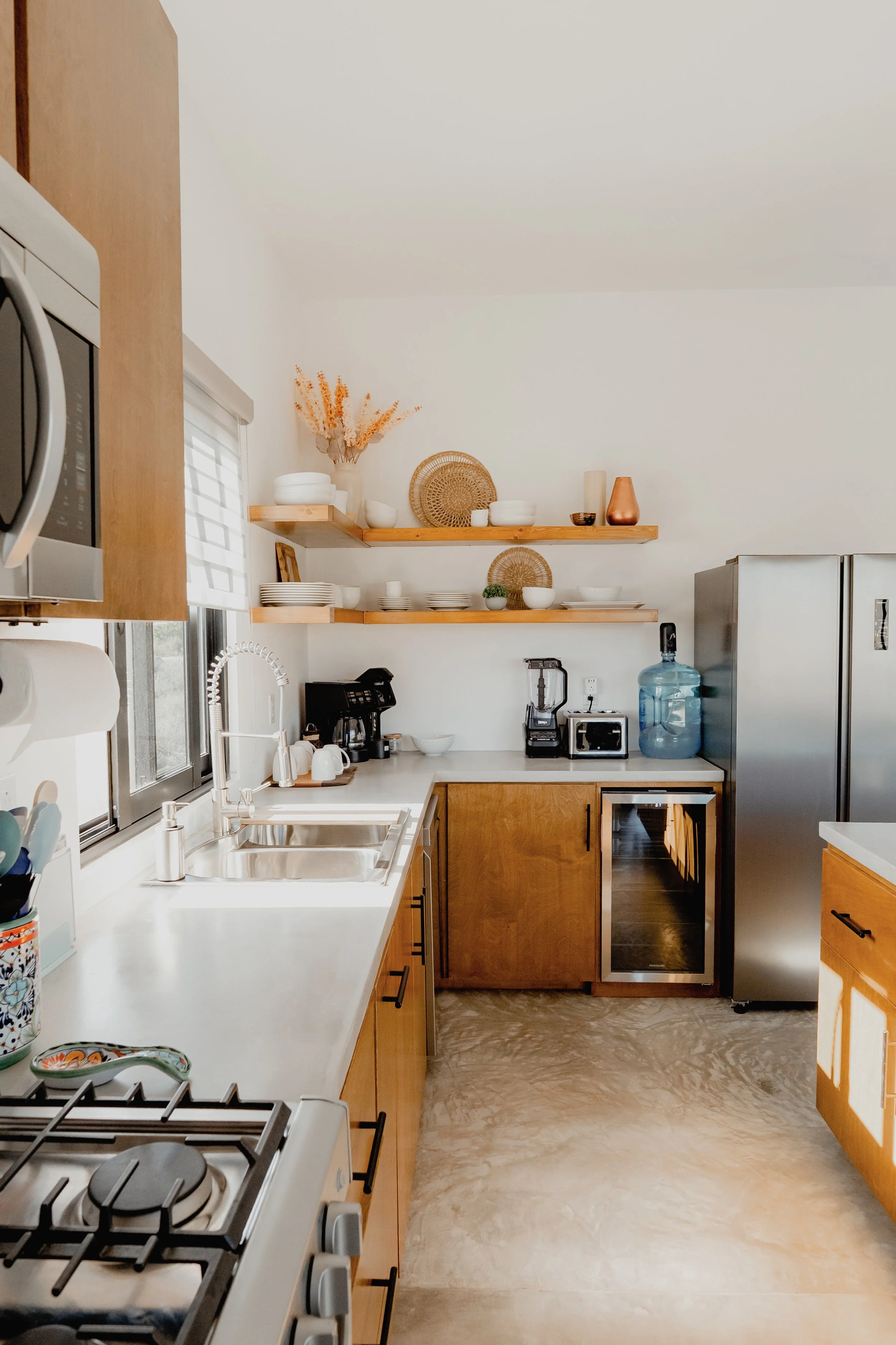 A kitchen with wooden cabinets, white countertops, open wooden shelves displaying bowls, plates, and decorative items, a stainless steel refrigerator, a water dispenser, and a black coffee machine on the counter near a window with blinds.
