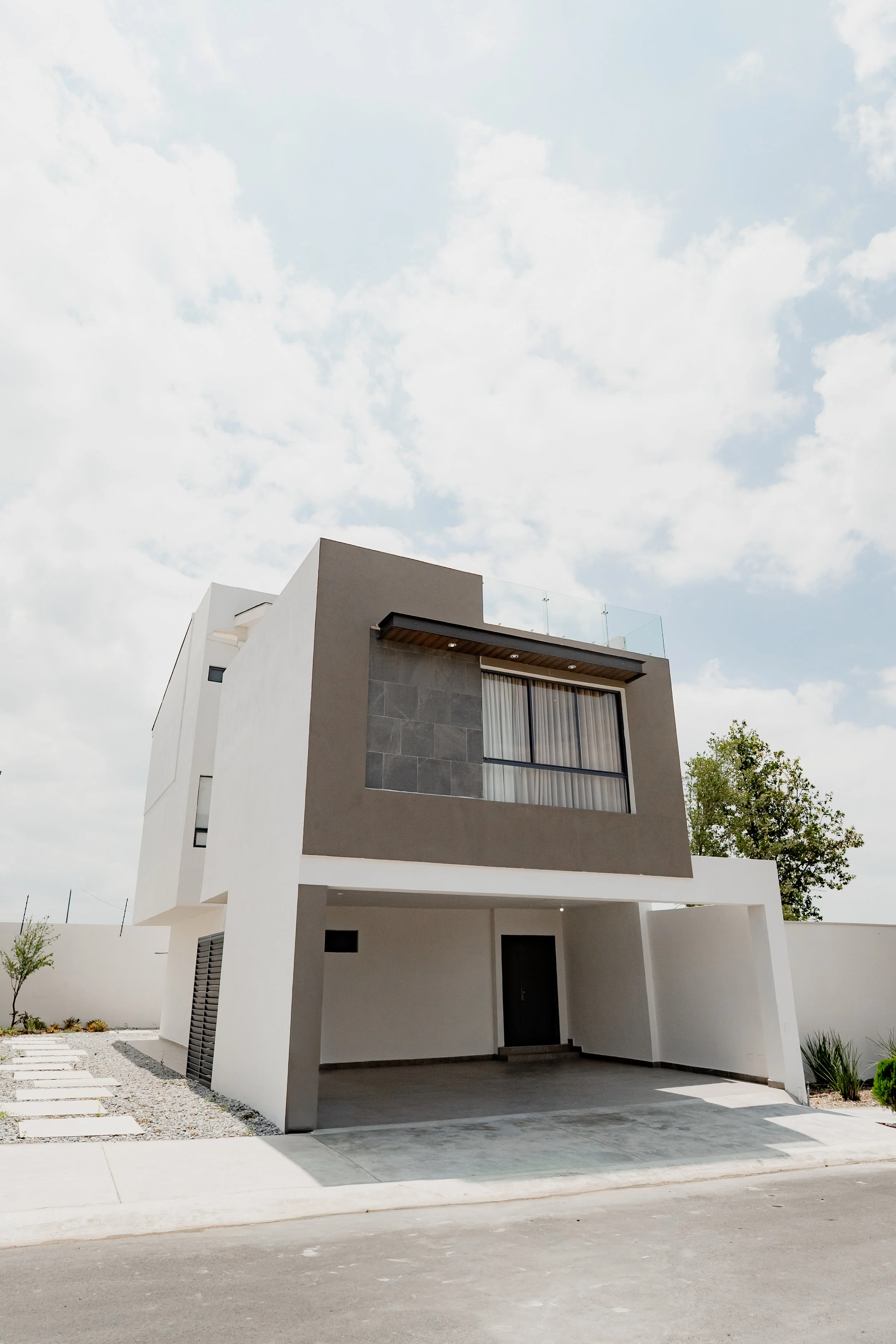 Modern white and gray house with a large balcony, open garage, minimal landscaping, and partly cloudy sky.
