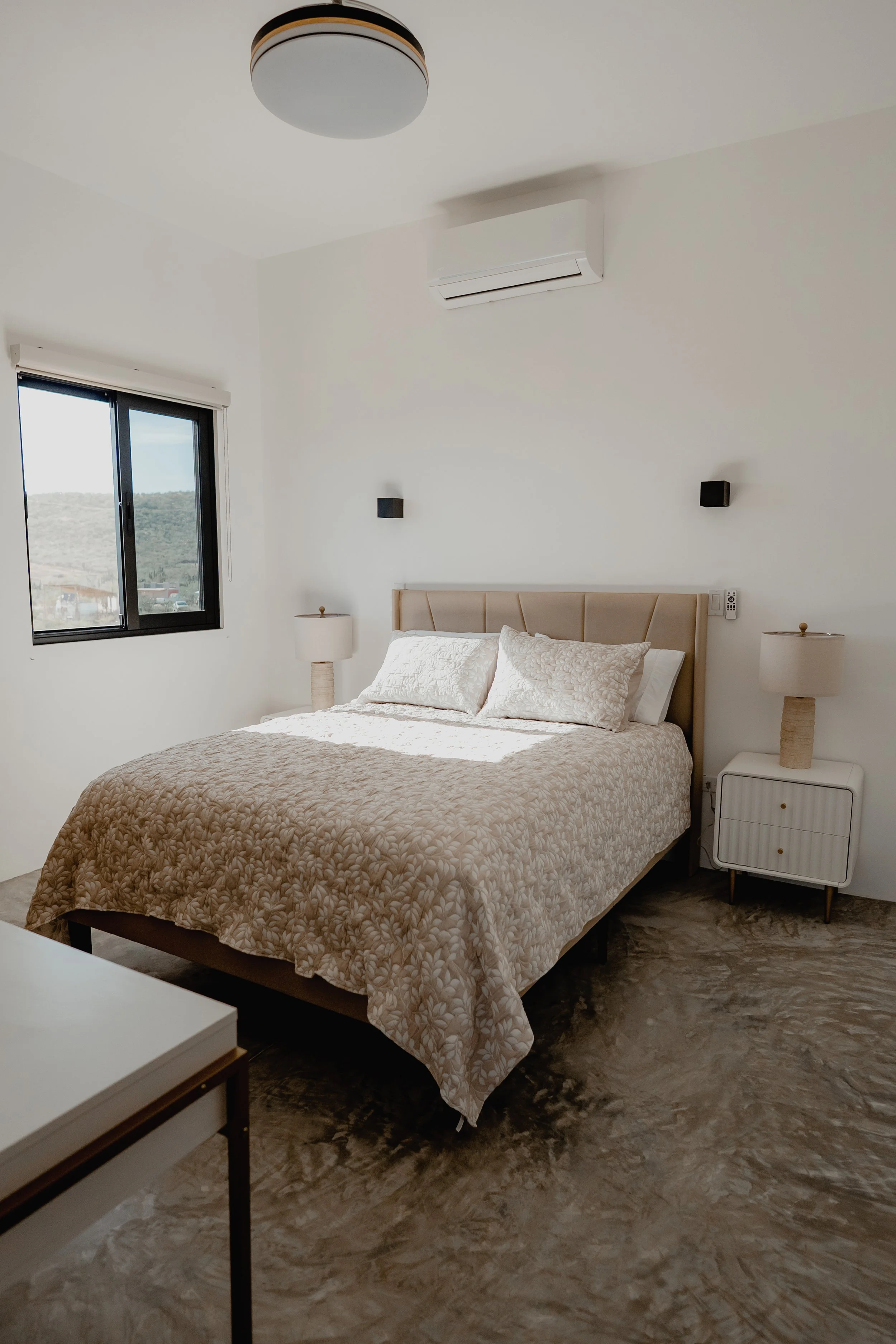 A bedroom with white walls, a bed with beige and white bedding, two nightstands with lamps, black wall-mounted speakers, a window with a mountain view, and a ceiling air conditioning unit.
