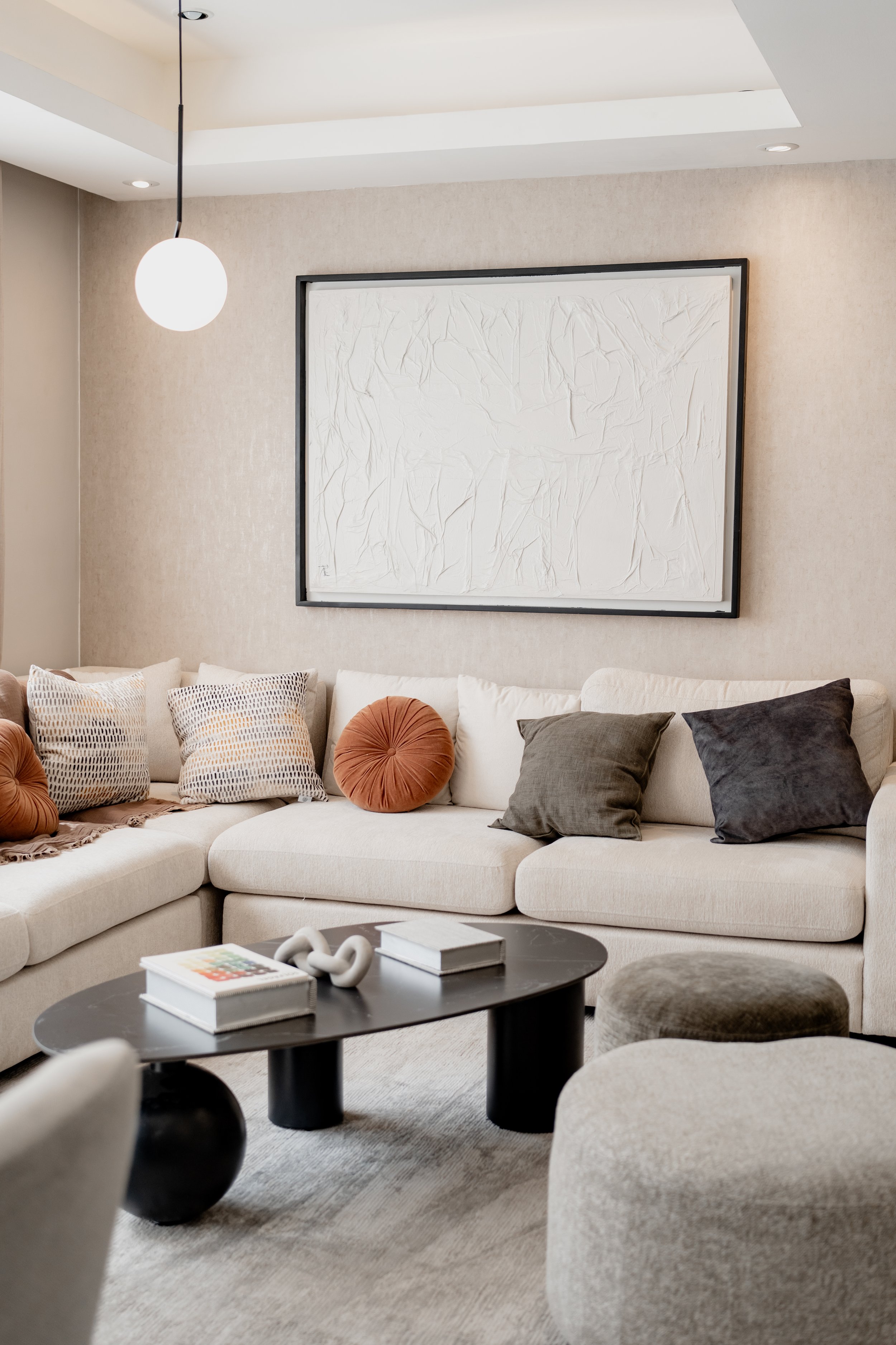 A living room with a beige sectional sofa, colorful throw pillows, a black coffee table with books and a decorative object, a gray ottoman, a textured beige rug, a large abstract white artwork on the wall, beige walls, a round hanging light, and ceil