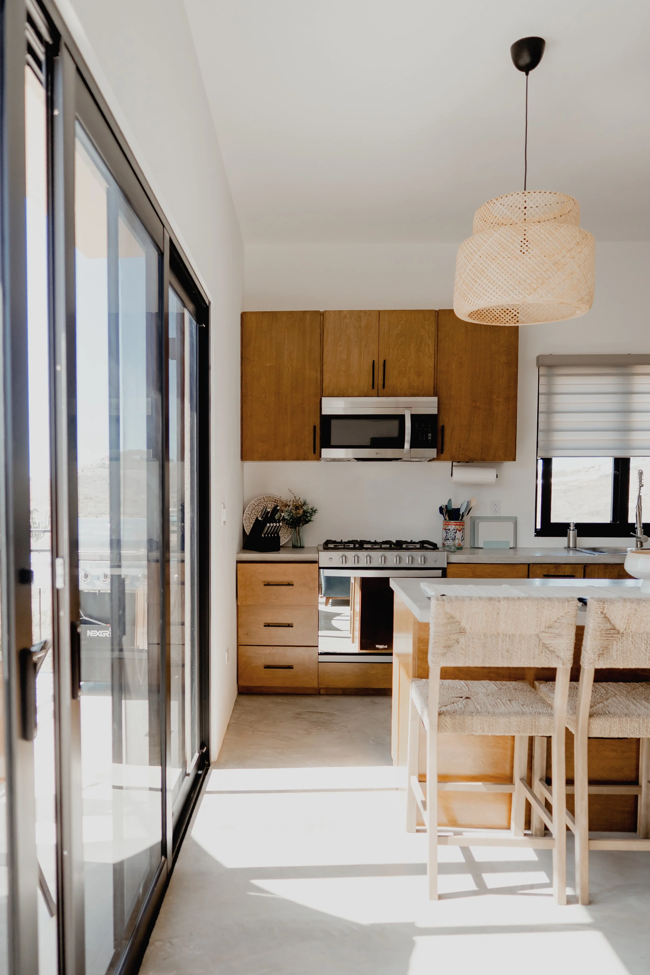A modern kitchen with wooden cabinets, a microwave oven, a gas stove, and white countertops. There are two chairs at a wooden dining table, a sliding glass door on the left, and a window above the sink on the right. The ceiling has a woven pendant li