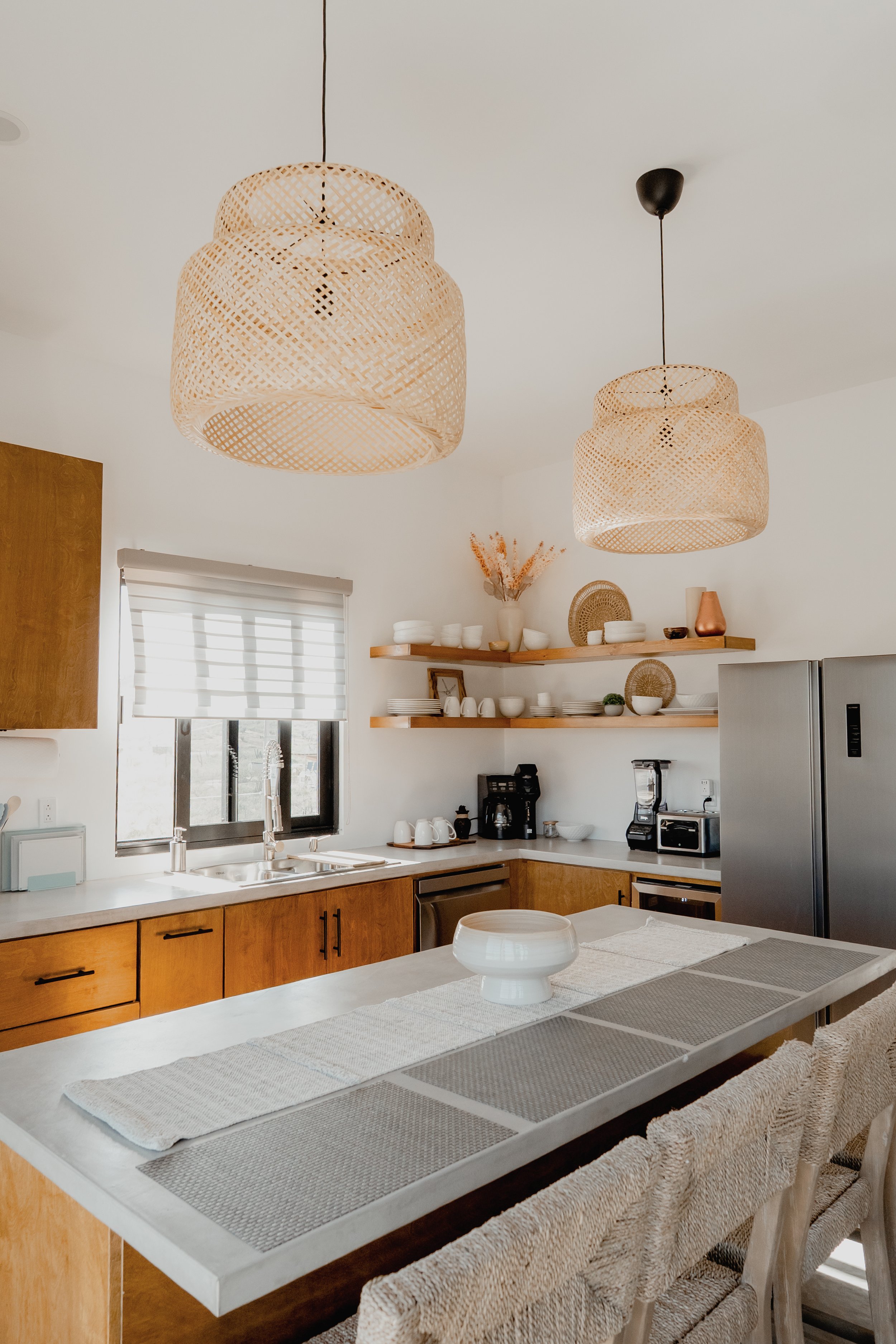 Modern kitchen with two wicker pendant lights hanging from the ceiling, wooden cabinets, open wooden shelves with white bowls and decorative items, a window with blinds, a countertop with small appliances, and a dining table with a white bowl centerp