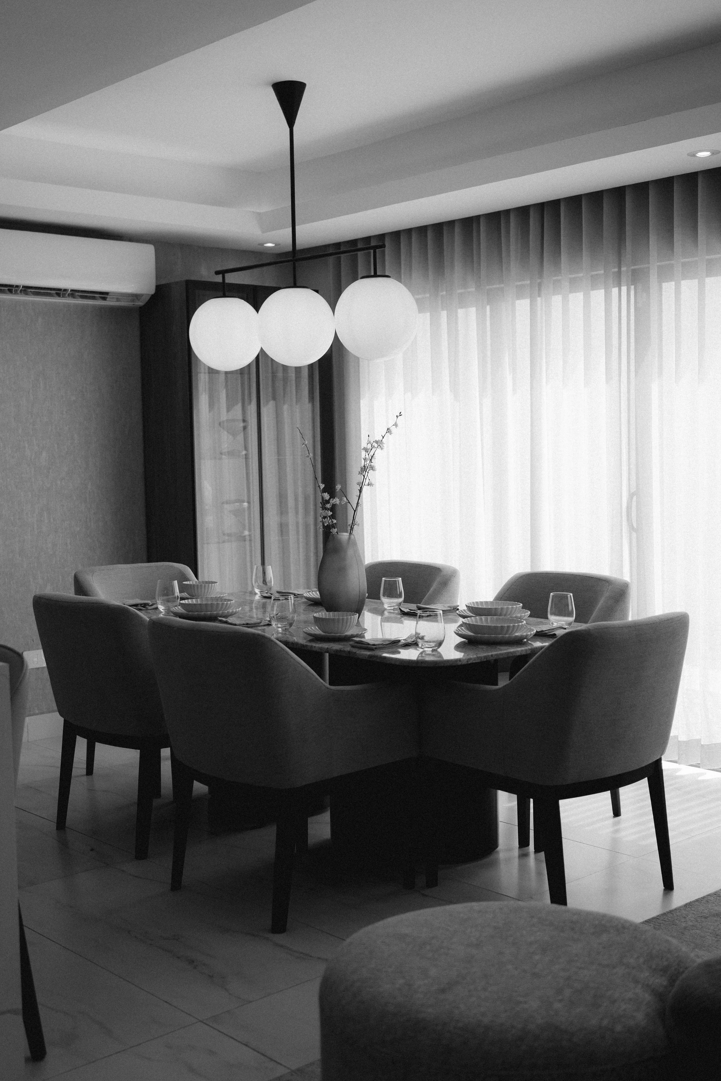 A dining room with a dark wood oval table set with bowls, glasses, and utensils, surrounded by cushioned chairs, a vase with branches in the center, large window with sheer curtains, and a hanging ceiling light fixture with three globes.