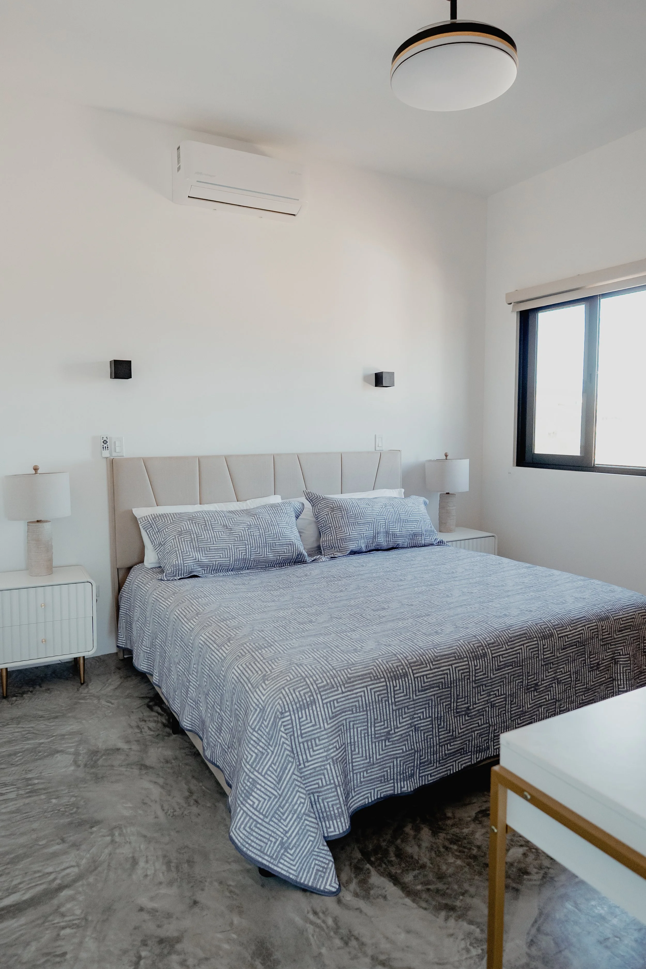 A modern bedroom with a bed, two nightstands with lamps, a window with a blind, a ceiling light, a wall air conditioner, and wall-mounted speakers.