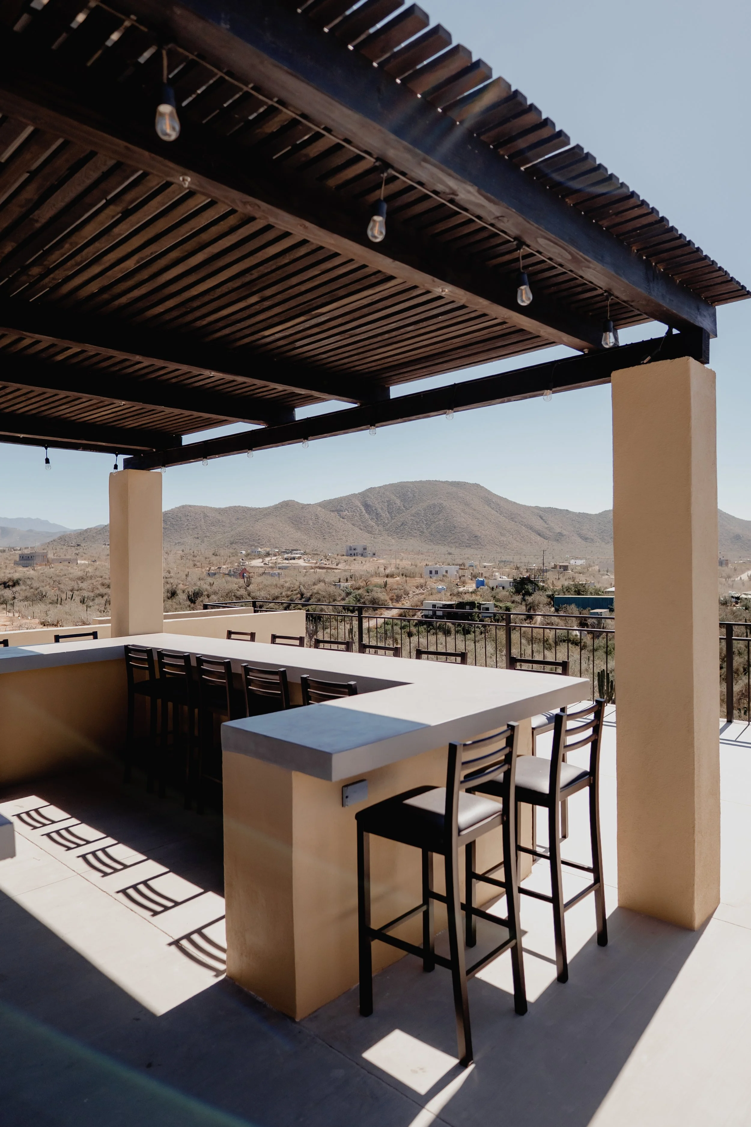Outdoor patio with high tables and chairs, shaded by a wooden pergola, overlooking a desert landscape with mountains in the background.