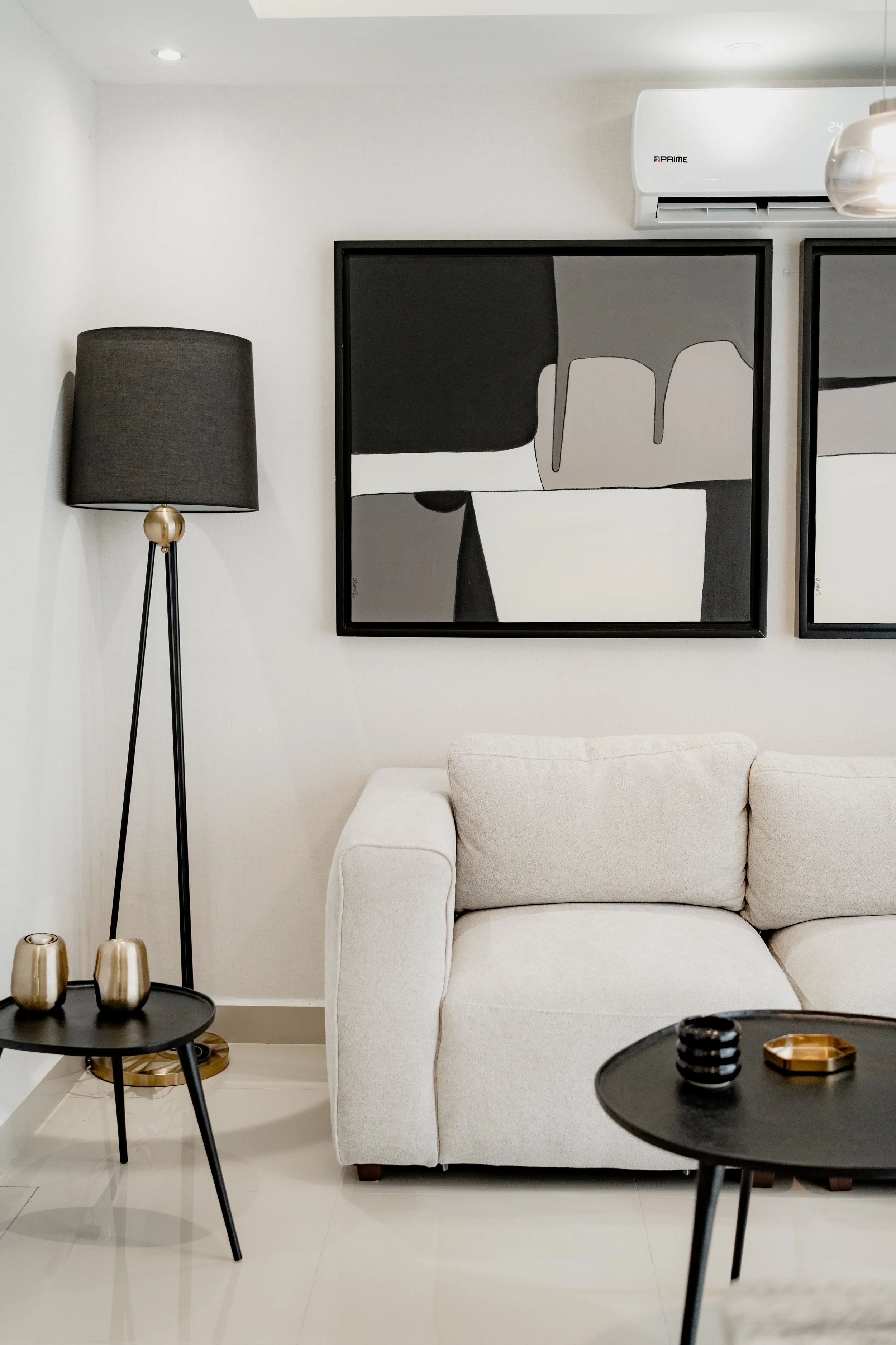 Living room with beige sofa, black side tables with gold decor, black lampshade, abstract black-and-white artwork on a white wall, and a white air conditioning unit.