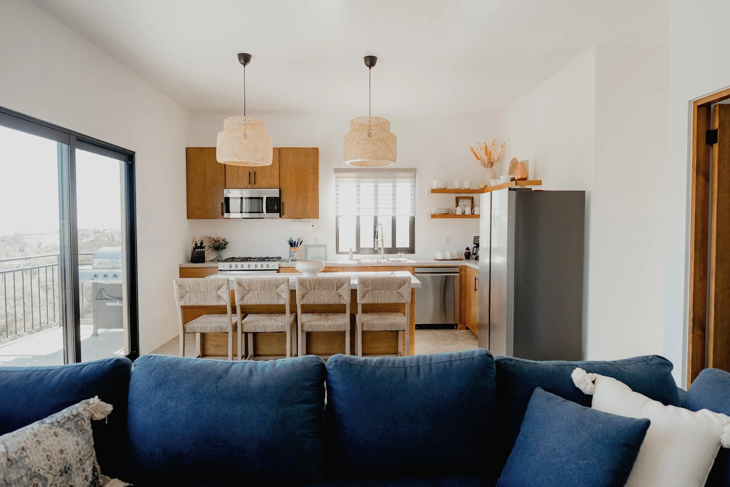 Open-concept living room and kitchen with a navy blue sofa, beige throw pillows, large glass sliding door, wooden dining table with beige chairs, kitchen with wooden cabinets, stainless steel appliances, and hanging woven pendant lights.