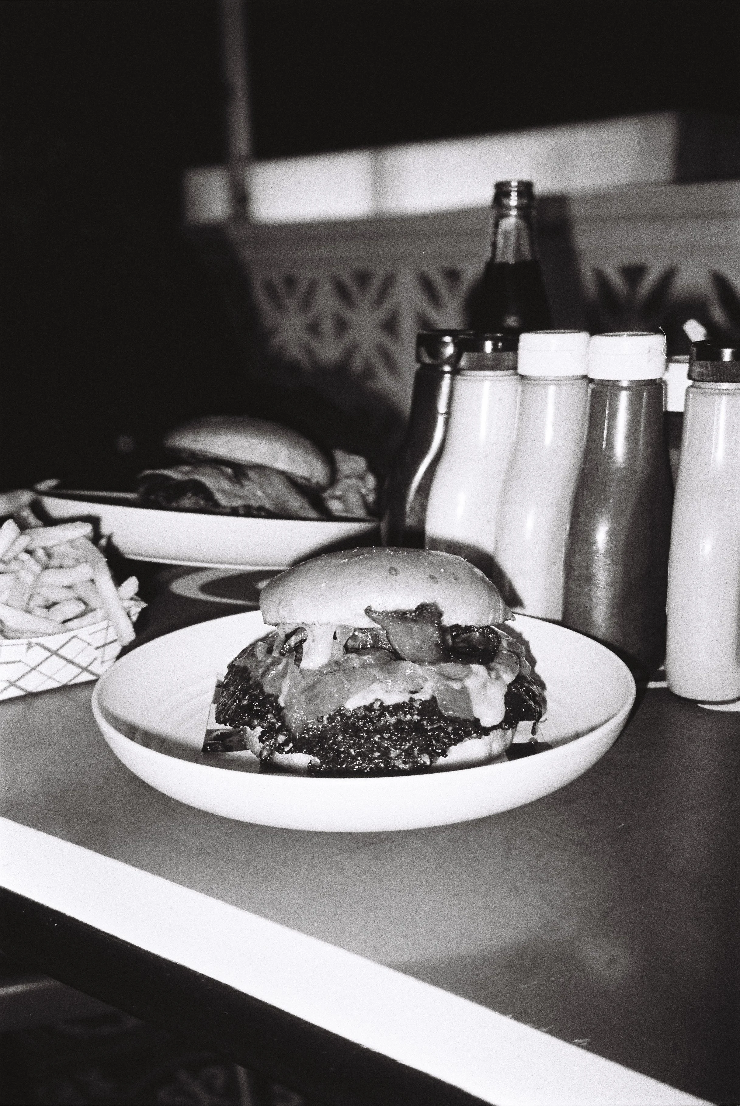 A burger with toppings and melted cheese on a white plate. In the background, there are condiment bottles and a burger with lettuce and tomato.