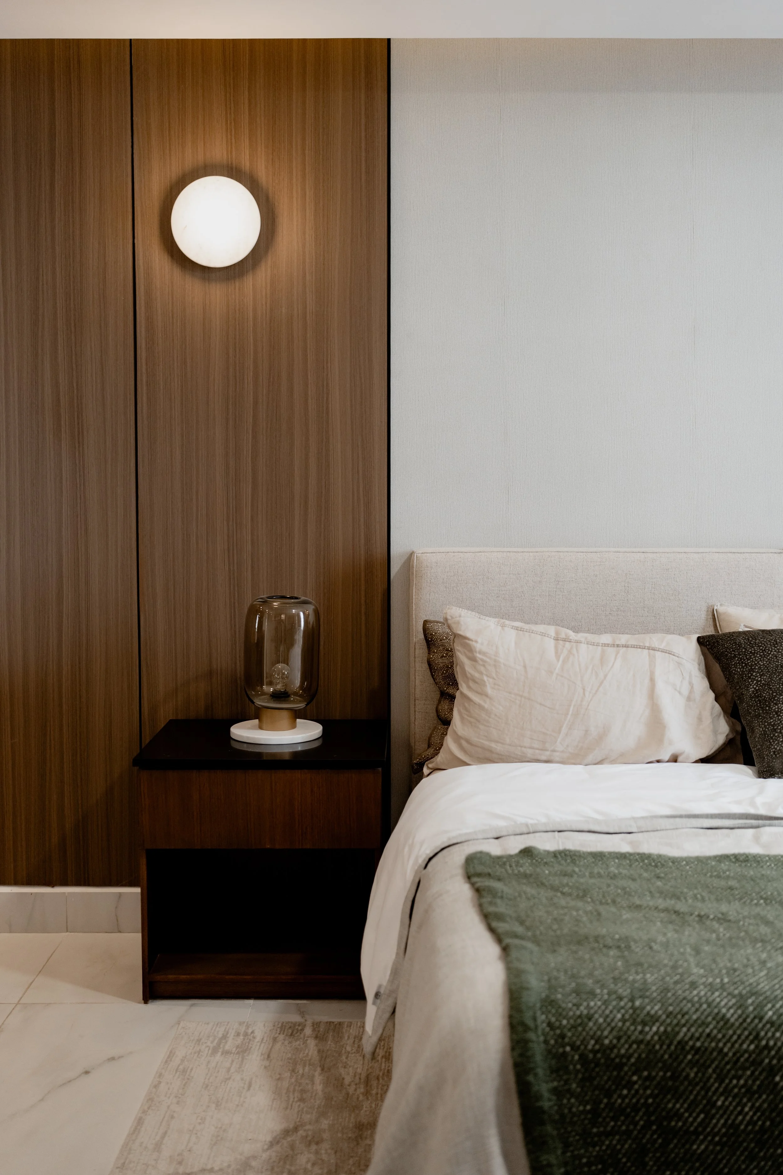 A bedroom with a modern wooden headboard, white pillow, beige bedding, green throw blanket, and a minimalist nightstand with a glass lamp. The wall behind has a round wall light, and the floor is tiled with a light-colored rug.