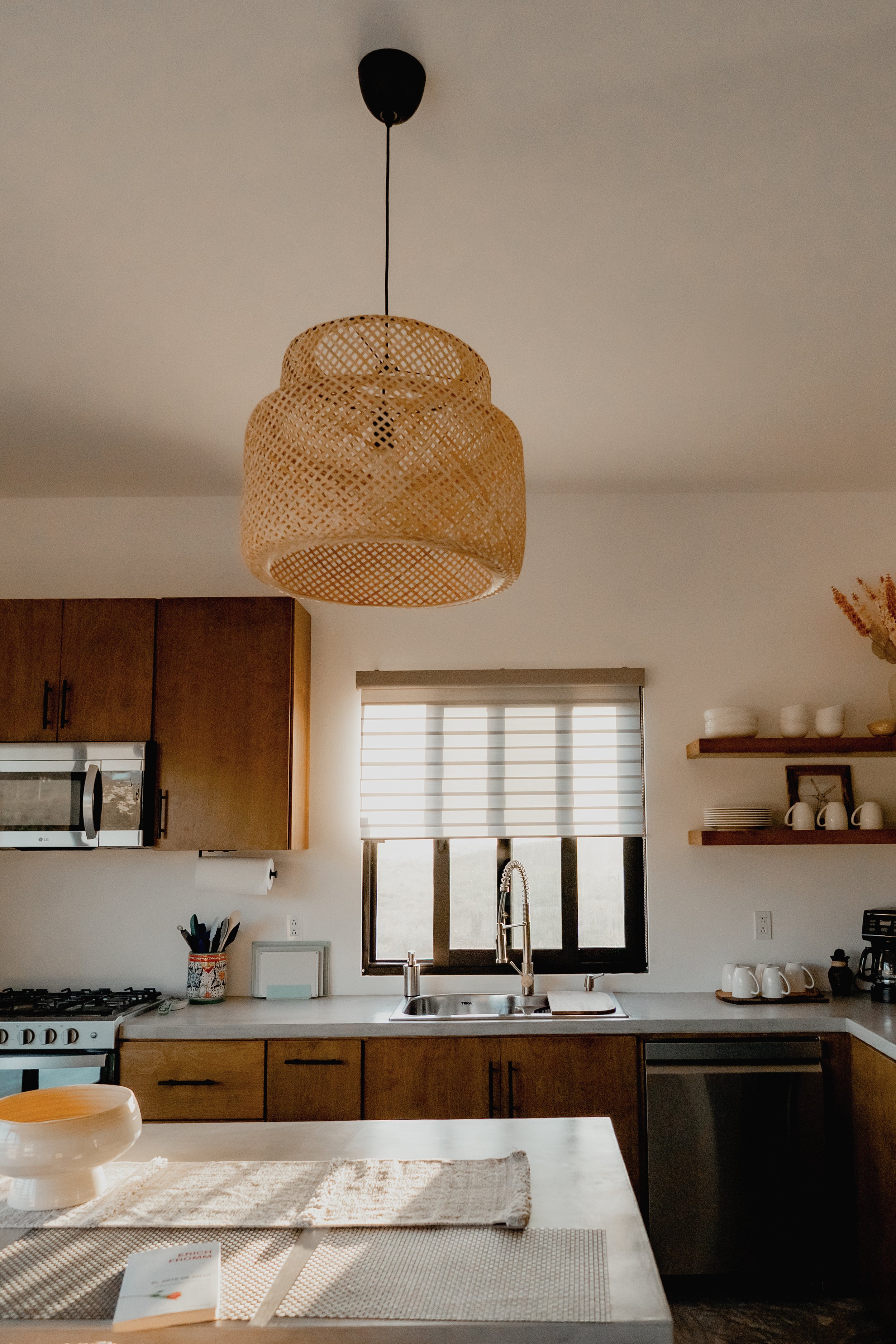 Kitchen with wooden cabinets, white countertop, stainless steel sink, window with blinds, hanging rattan light fixture, stove, and various dishware on shelves and counter.