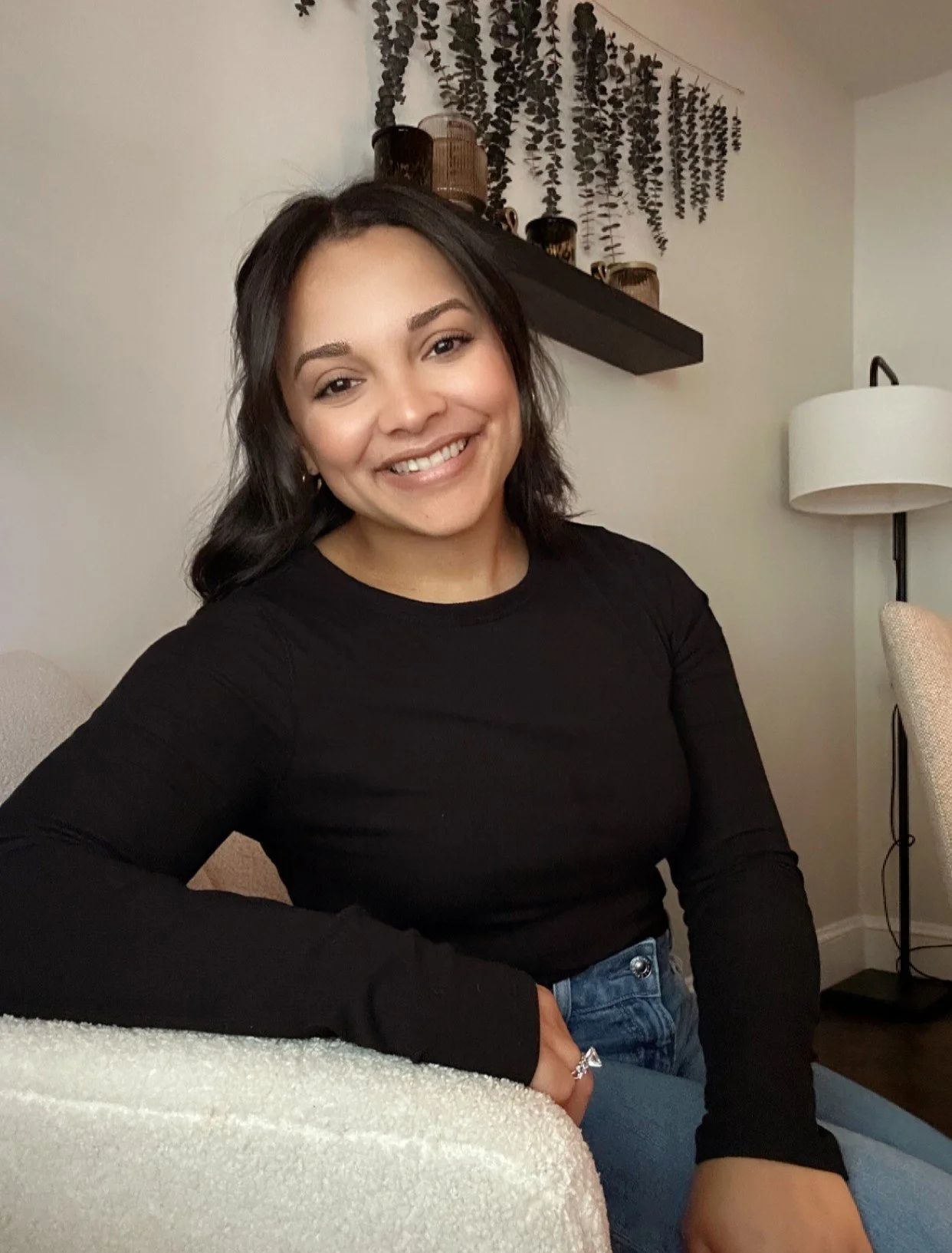 A young woman with shoulder-length dark hair, smiling, wearing a black long-sleeve shirt, sitting in a cozy room with a beige wall, a floor lamp, a wall shelf with decorative vases, and hanging greenery.
