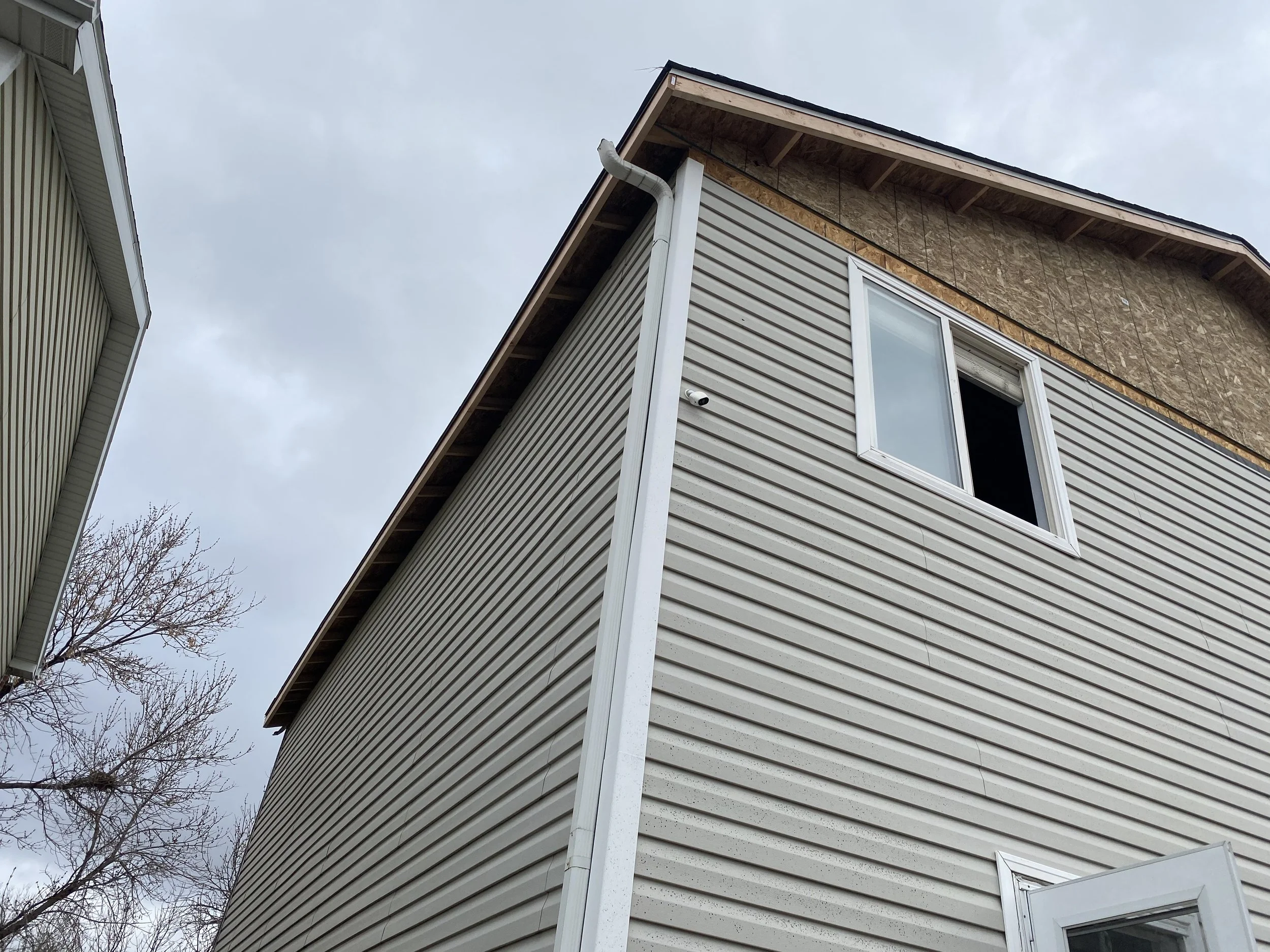 Side of a house with vinyl siding, featuring vertical, diagonal, and horizontal patterns, with some bushes on the right side.