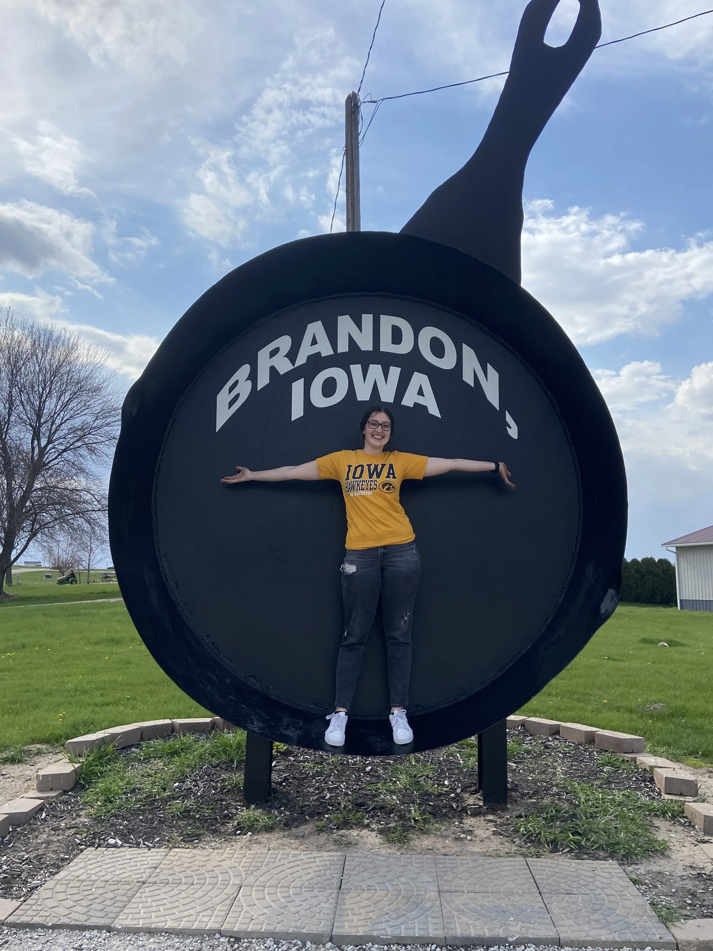 A woman standing with arms outstretched inside a large black frying pan sculpture in BRANDON IOWA on the side.