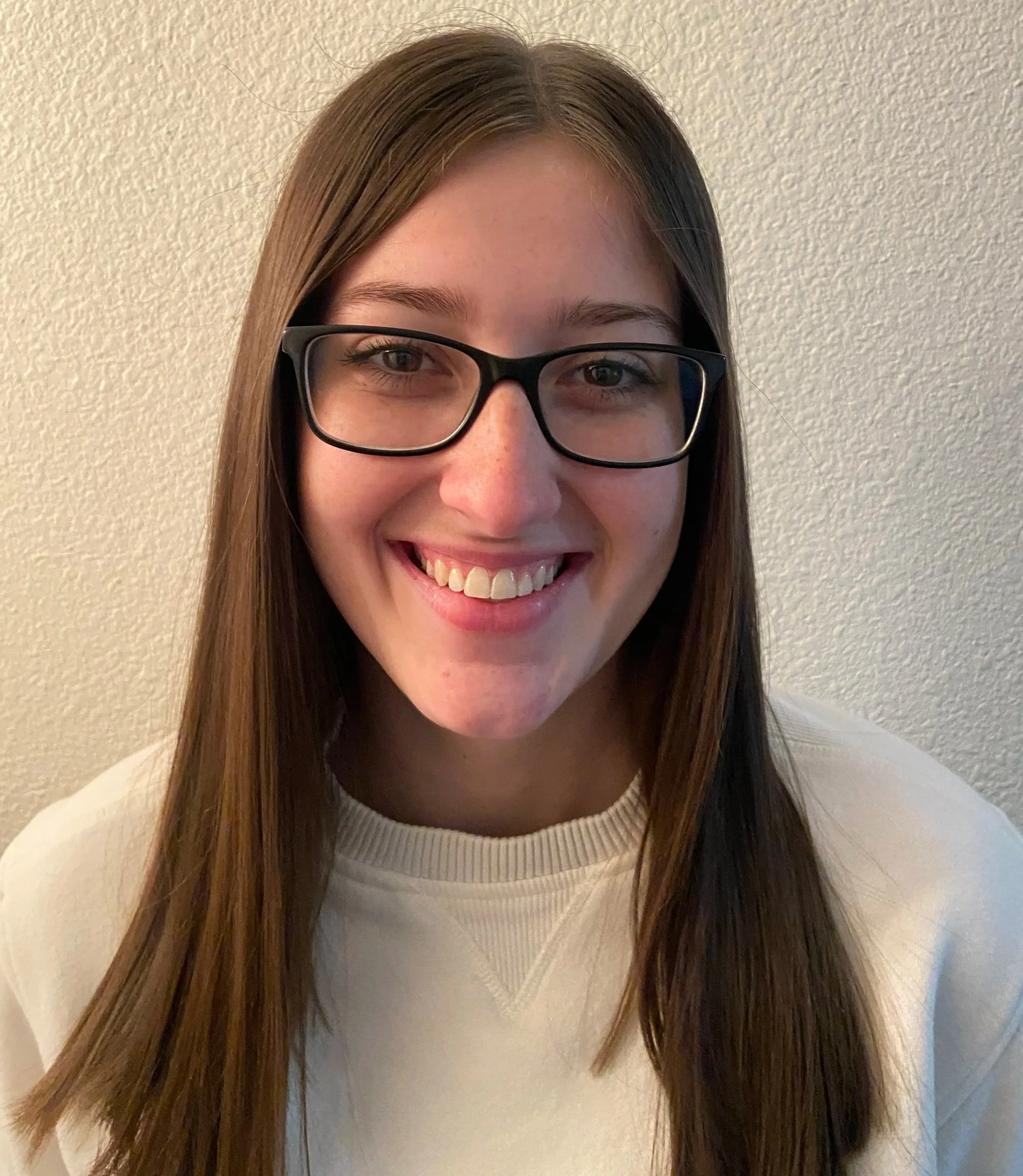 A young woman with long brown hair, wearing glasses and a white sweatshirt, smiling at the camera against a textured beige wall.
