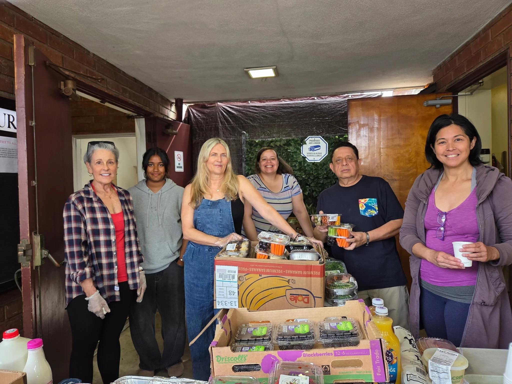 Group of six people standing behind a table with food supplies in an indoor community setting, smiling at the camera.