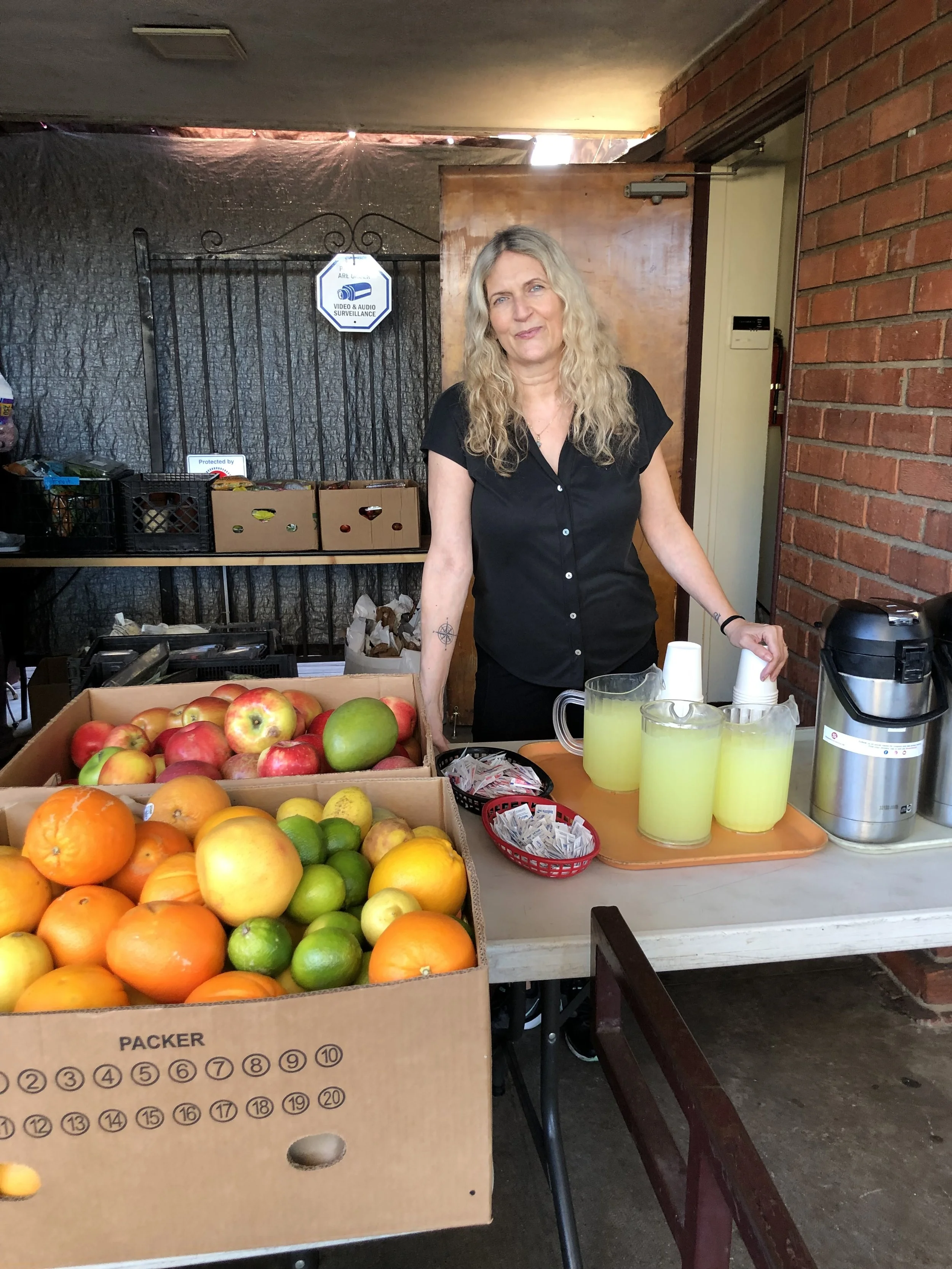 Lisa standing behind a table with fresh fruit and juice pitchers, in a setting with brick walls and a black wire fence in the background.