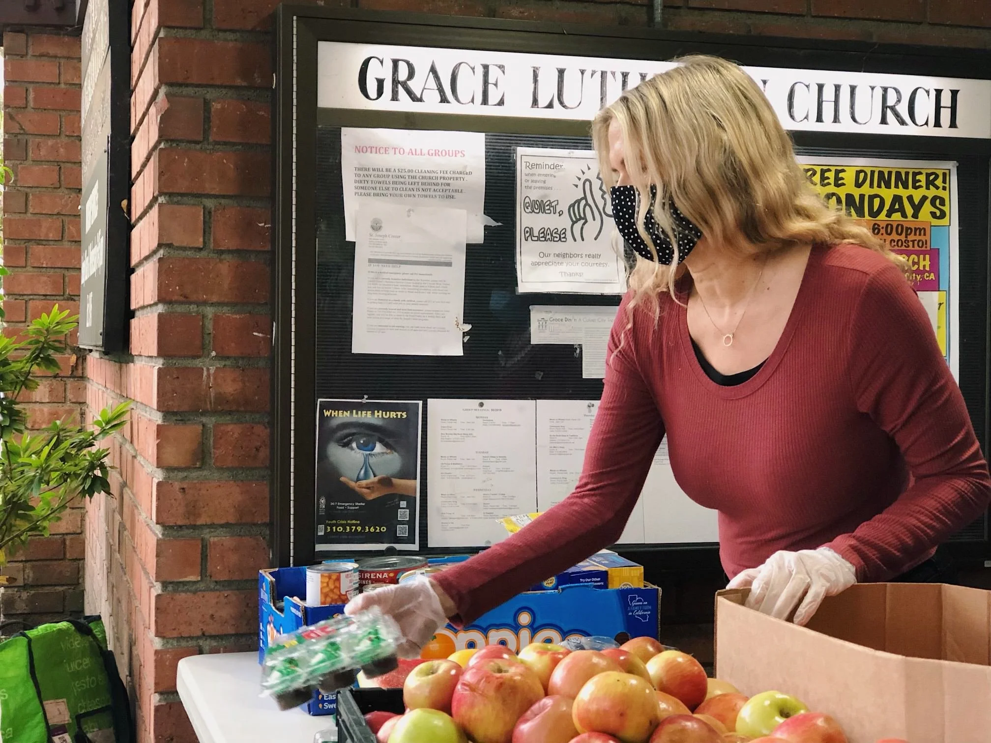 Lisa Skelley, wearing a black face mask with white polka dots, is organizing food donations at Grace Diner, with a box of apples and canned goods on the table in front of her. The background shows bulletin boards and notices outside the church.