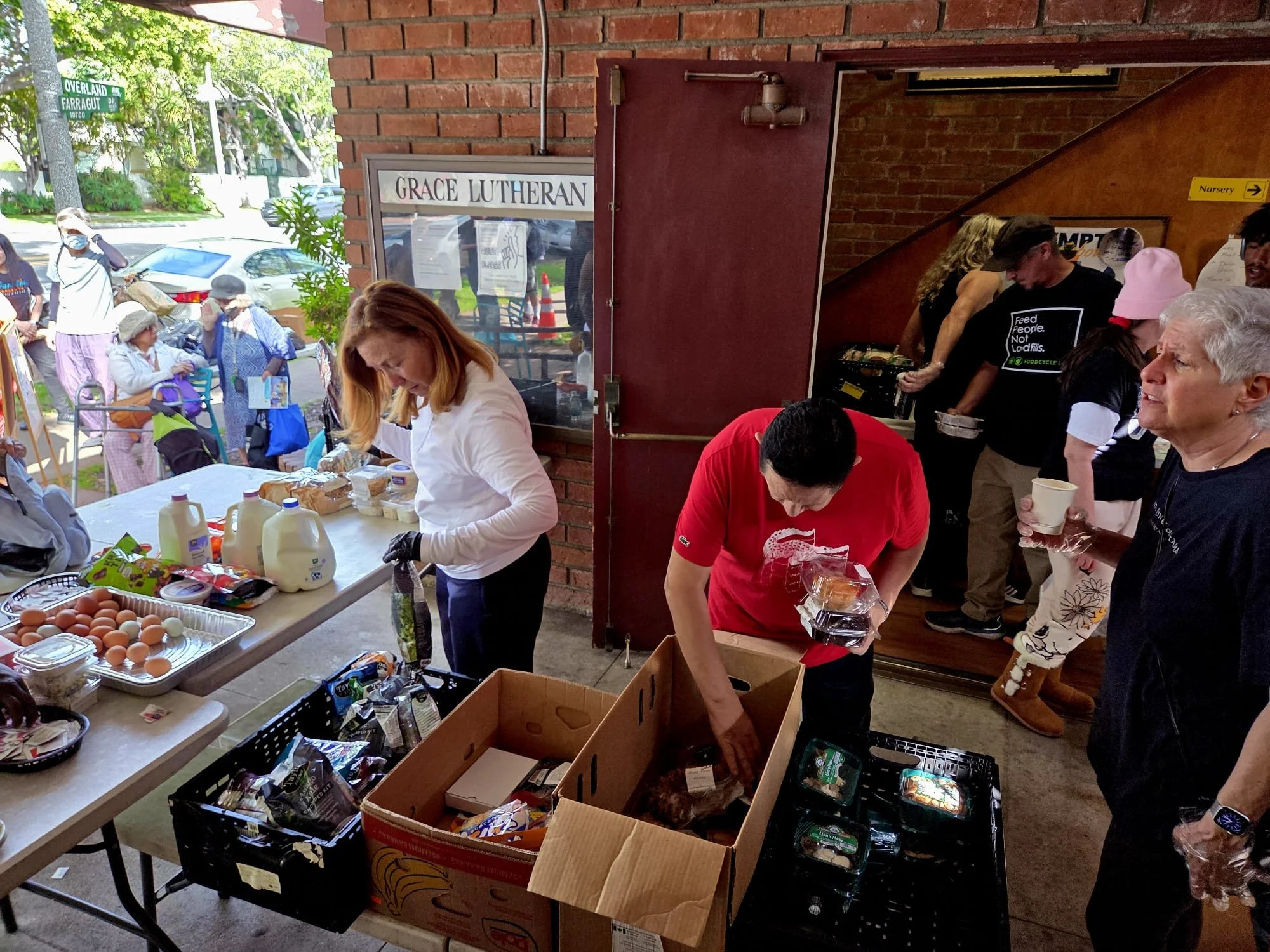 People volunteering to prepare and distribute food and supplies at a community event outside a brick building.