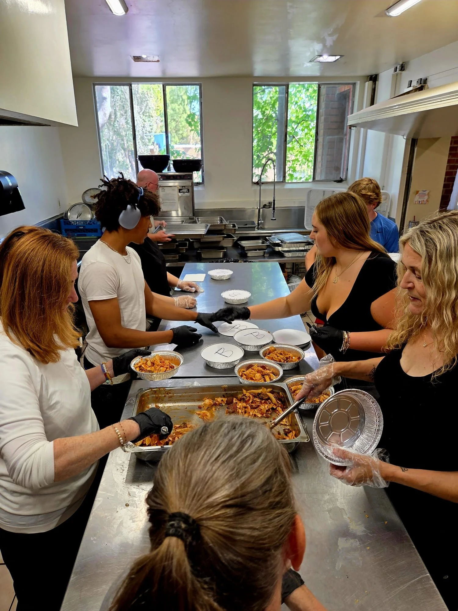 People in a kitchen serving food, with plates being filled from a tray of pasta, during a food distribution event.