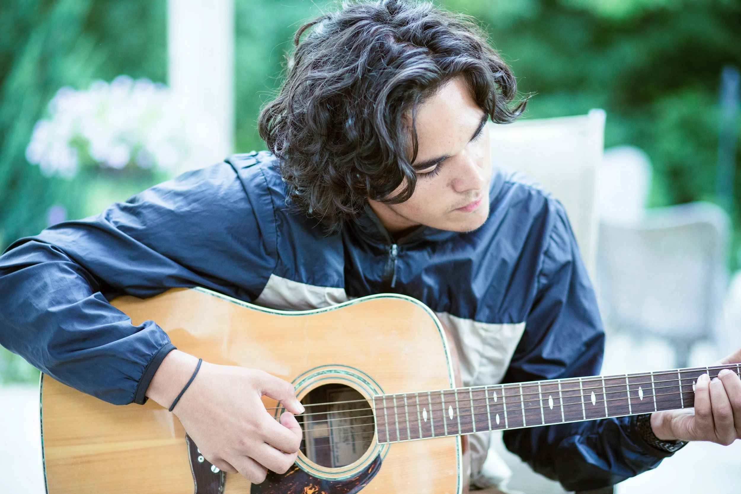 A man with curly dark hair playing an acoustic guitar outdoors, wearing a dark jacket, with a blurred green natural background.
