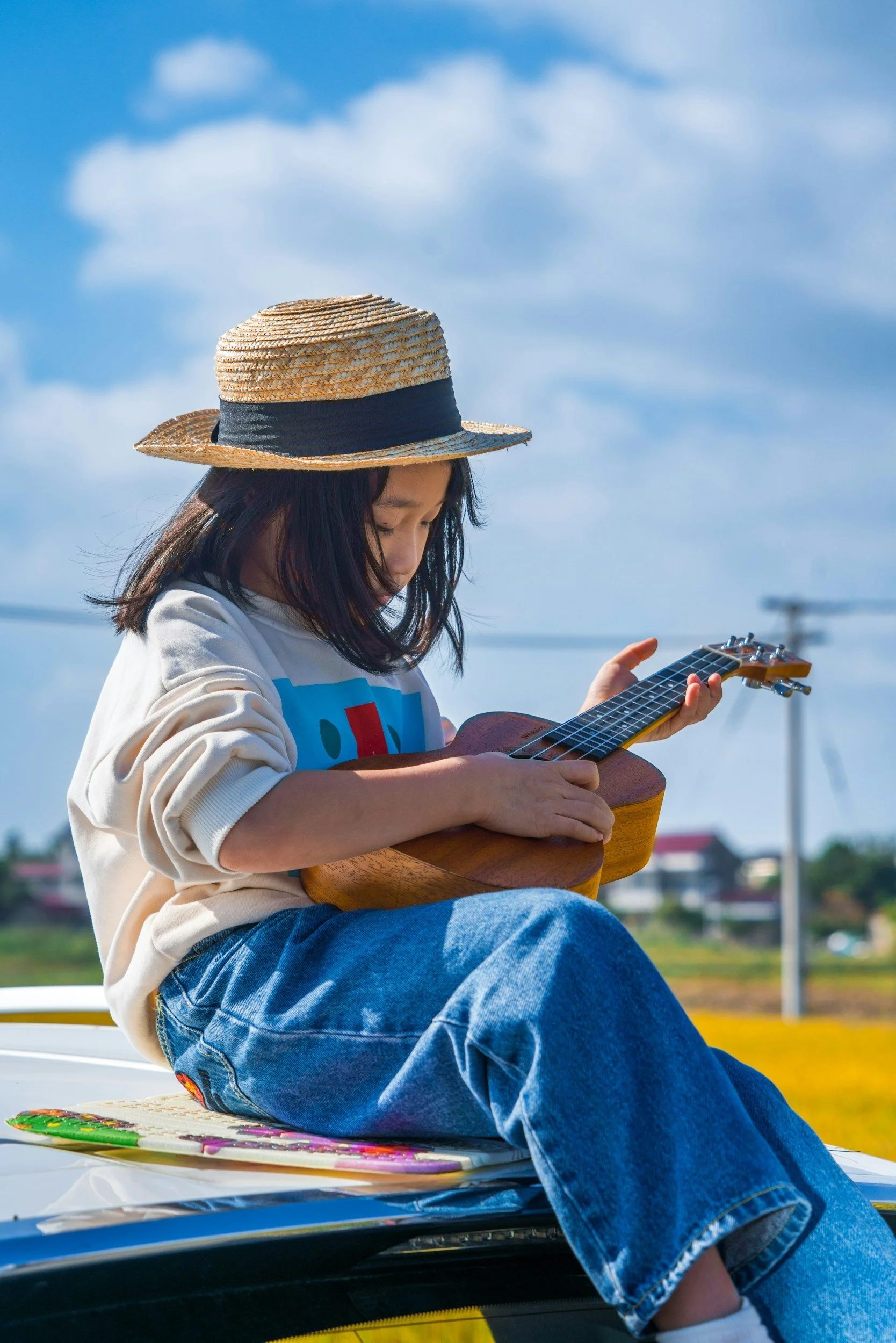 A music student wearing a straw hat playing an acoustic guitar while sitting outdoors on a sunny day.