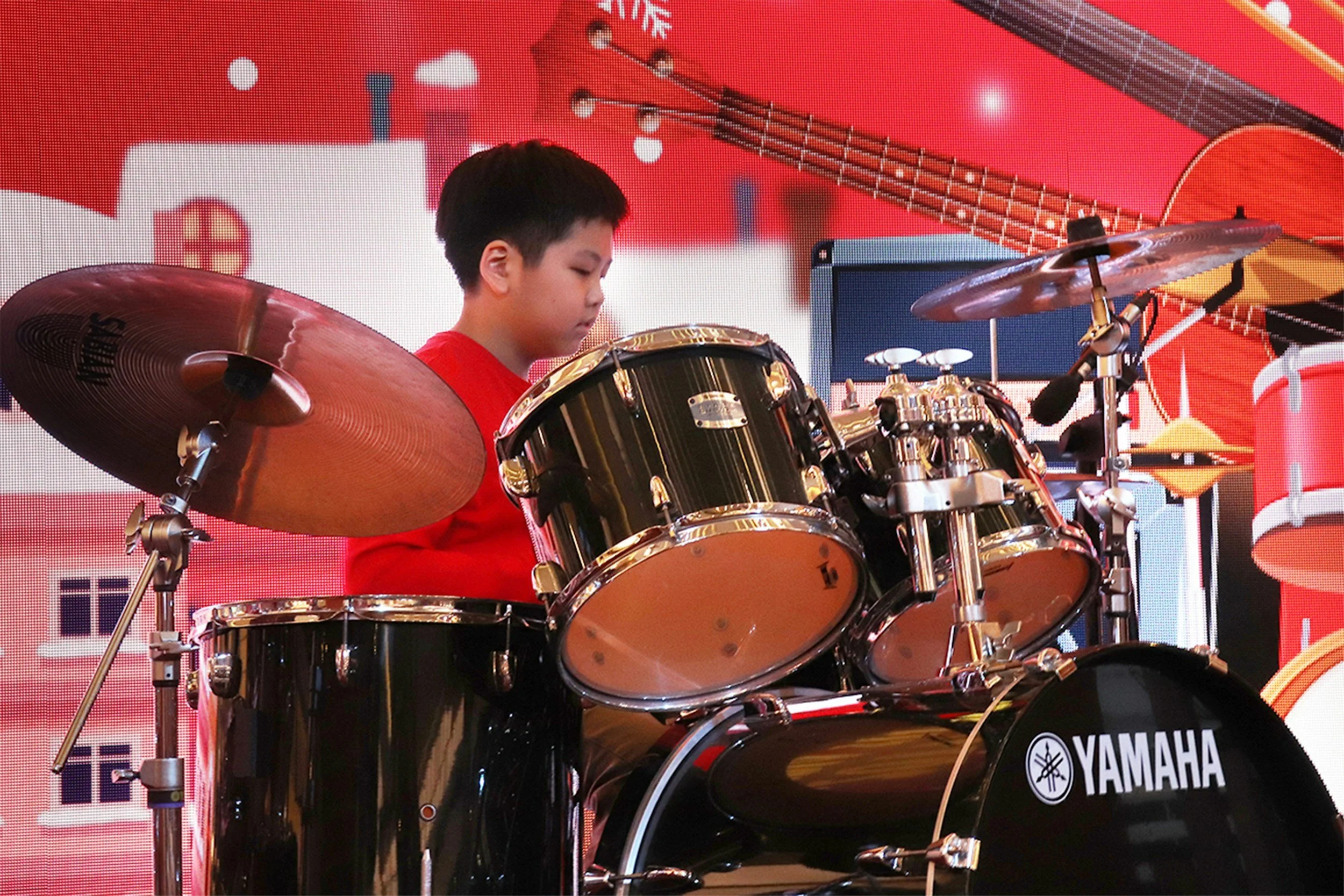 A young boy with short black hair playing a black Yamaha drum set with gold hardware in front of a red background with musical and Christmas-themed decorations.