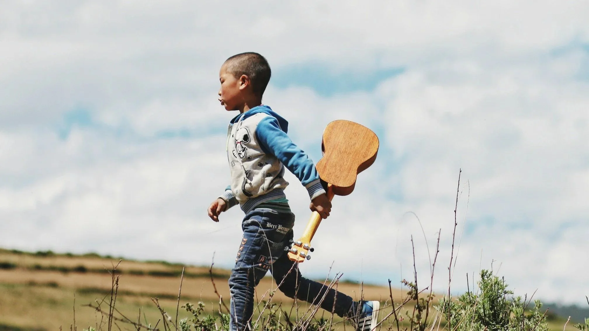 A young music student holding an acoustic guitar in his right hand, with a partly cloudy sky in the background.