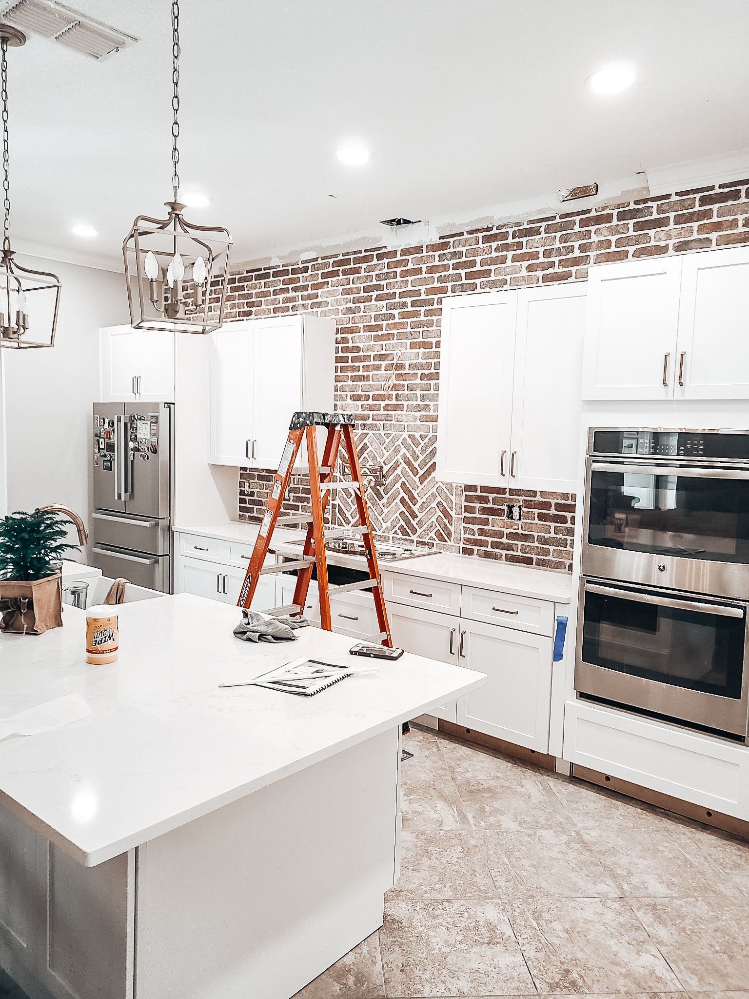 Kitchen with white cabinets, stainless steel appliances, brick wall, island, orange ladder, and the wall behind the stove in the process of being tiled.