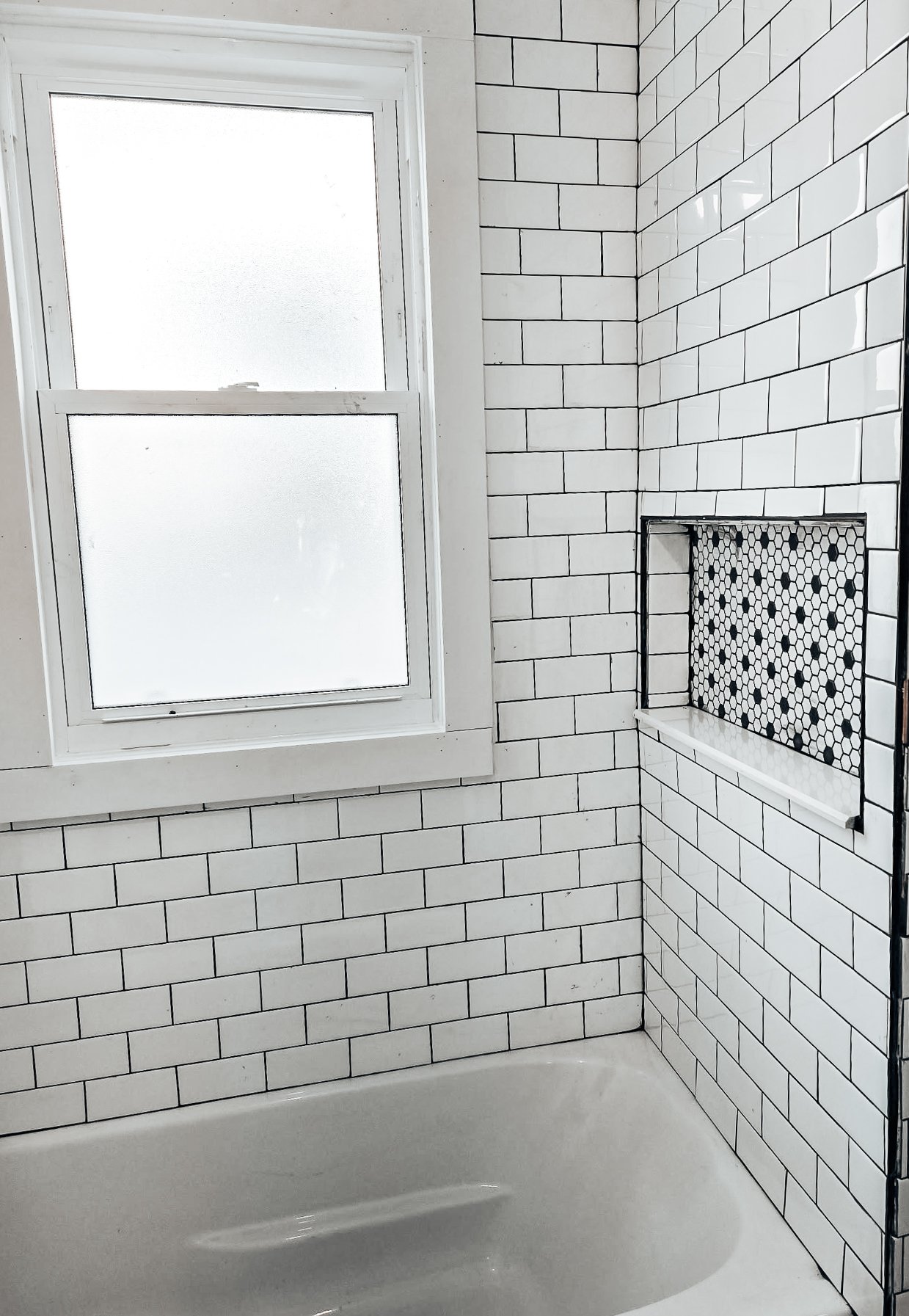 Bathroom with white subway tile walls, frosted window, and a small built-in shower niche with black and white mosaic tiles.
