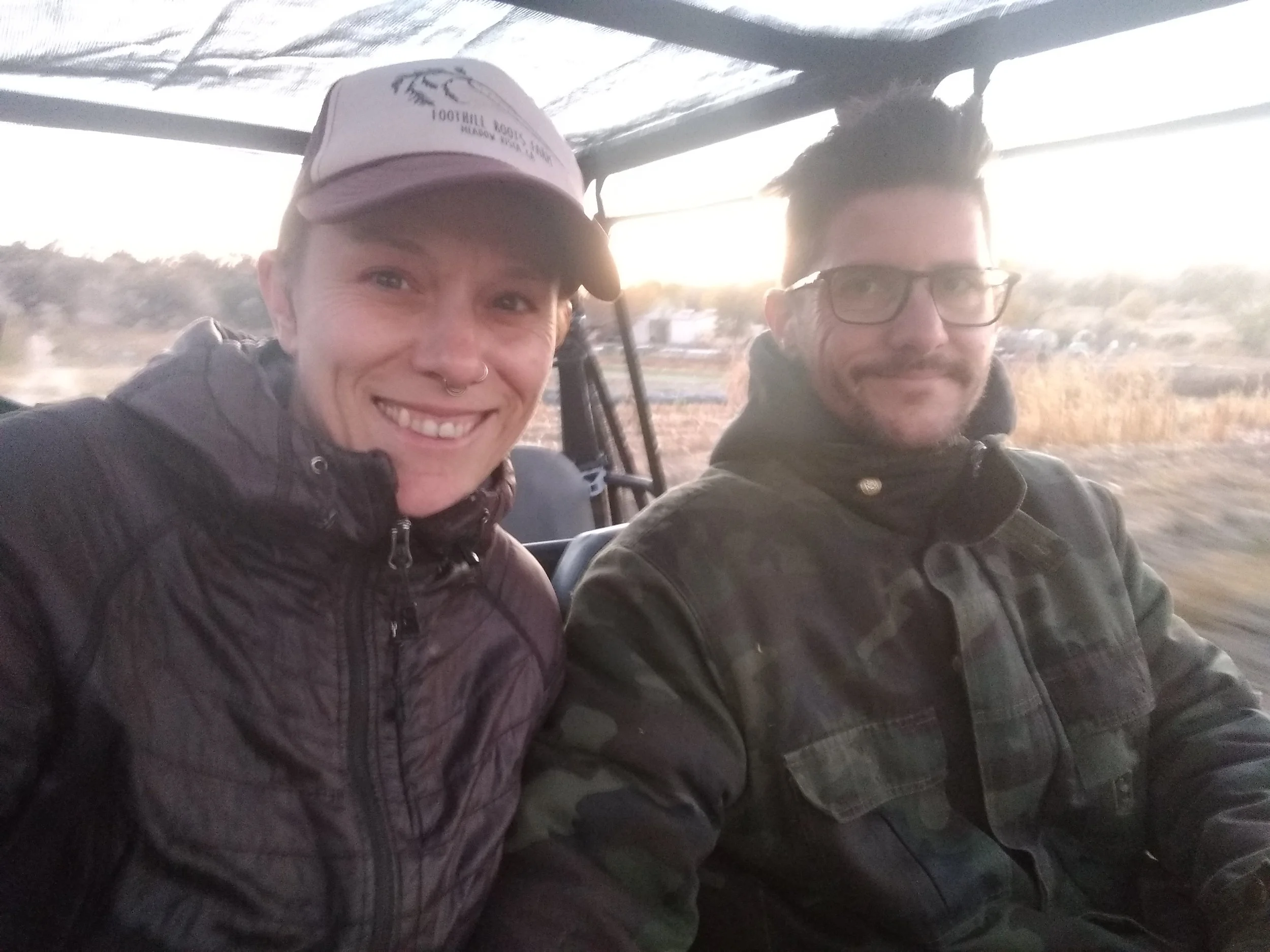 Two people sitting in a vehicle with a blurred outdoor background during sunset. The person on the left is smiling, wearing a cap and a black jacket. The person on the right wears glasses, a dark jacket, and has dark hair.