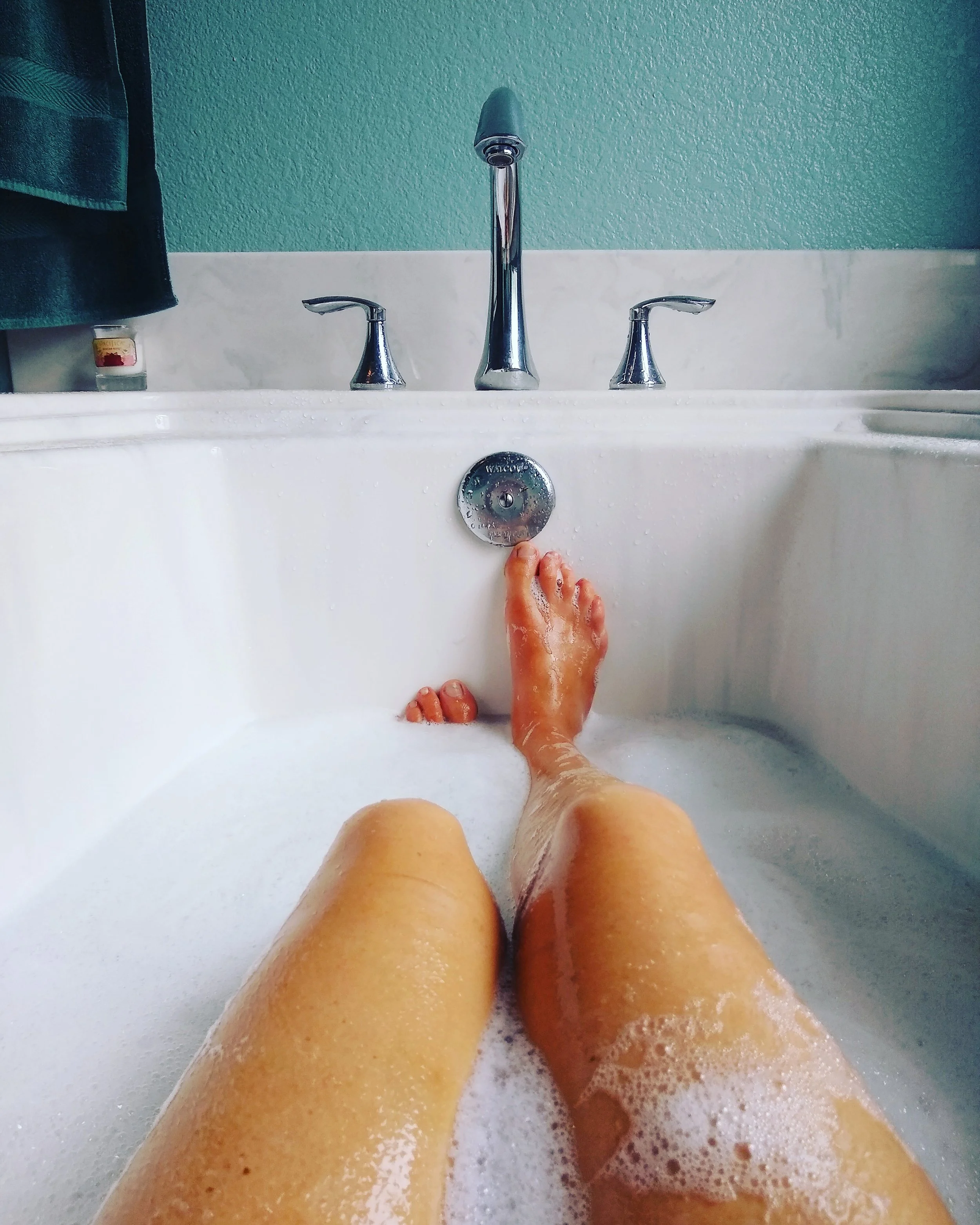 Person relaxing in a bathtub filled with bubbles, with legs visible and feet near the drain, bathroom walls visible in the background.