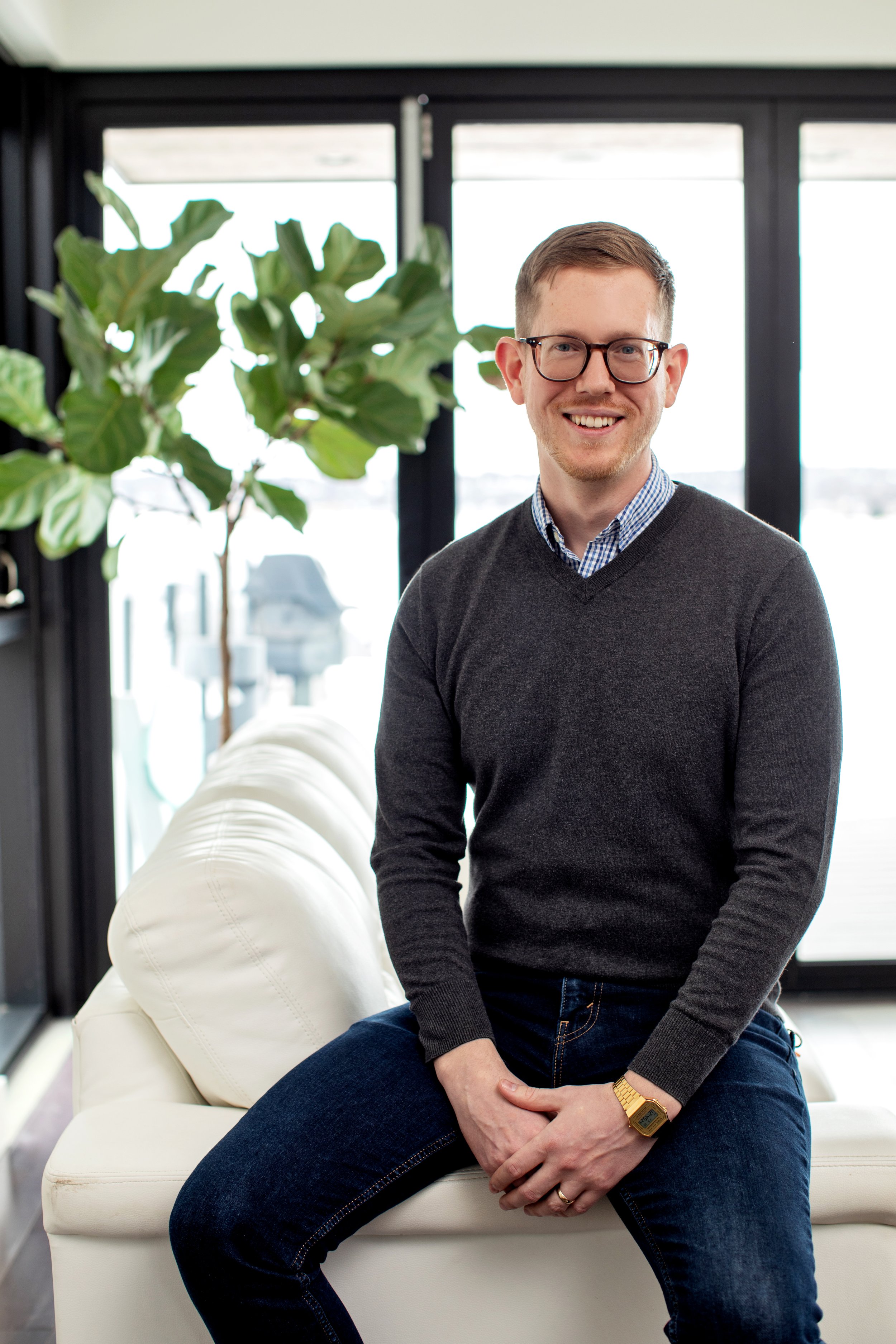 A man sitting on a white sofa in front of large windows, smiling, wearing glasses, a dark sweater over a blue checkered shirt, and jeans, with a large green plant in the background.