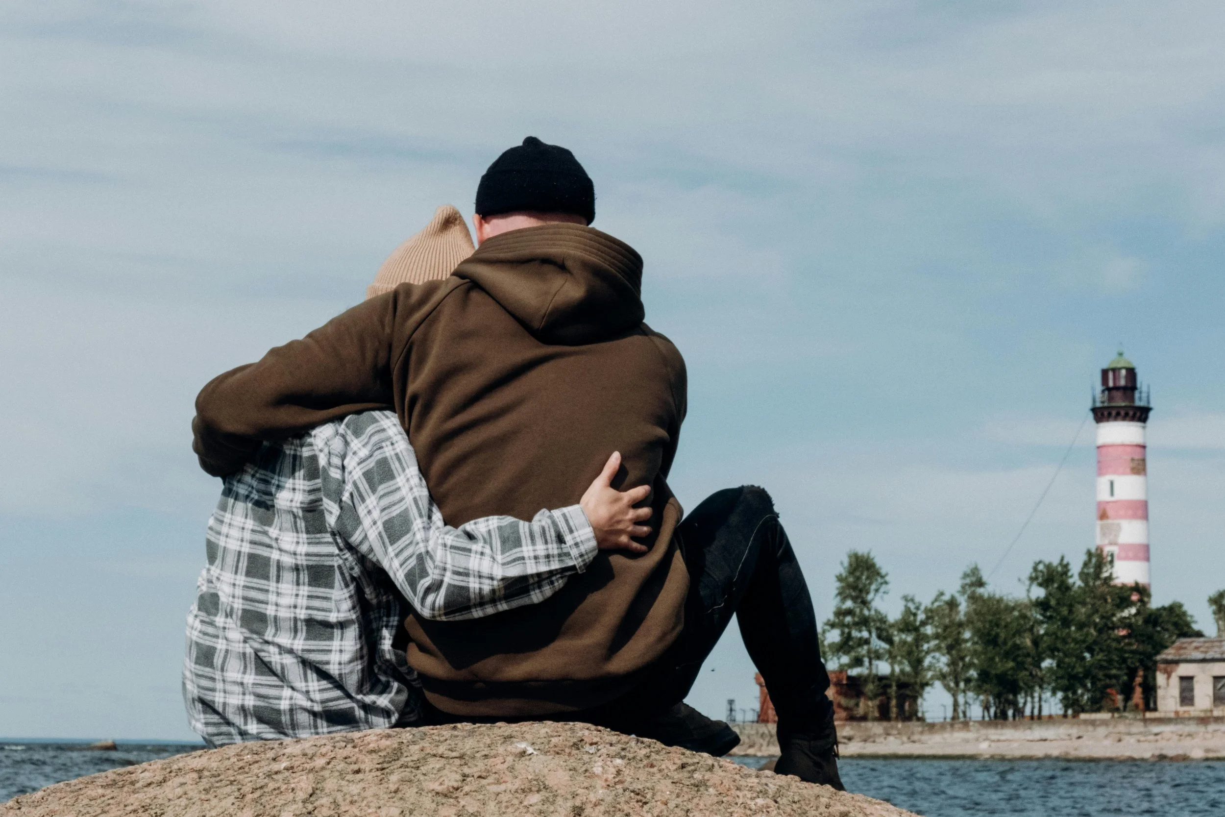 Two people sitting on a large rock by the water, embracing each other, with a lighthouse and trees in the background.