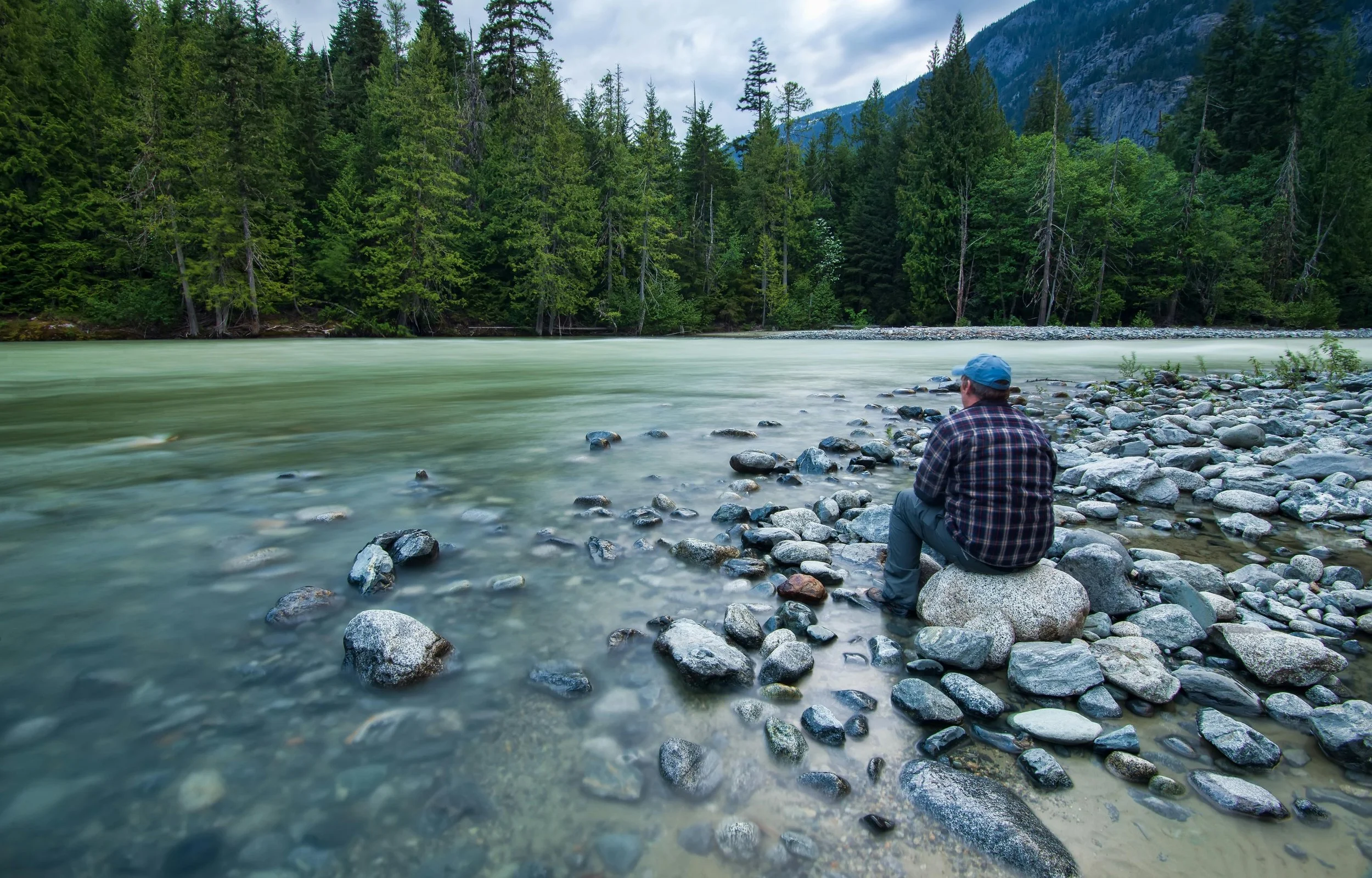 A man sits on a large rock by the riverbank, surrounded by smaller rocks, overlooking a flowing river with a forest and mountains in the background.