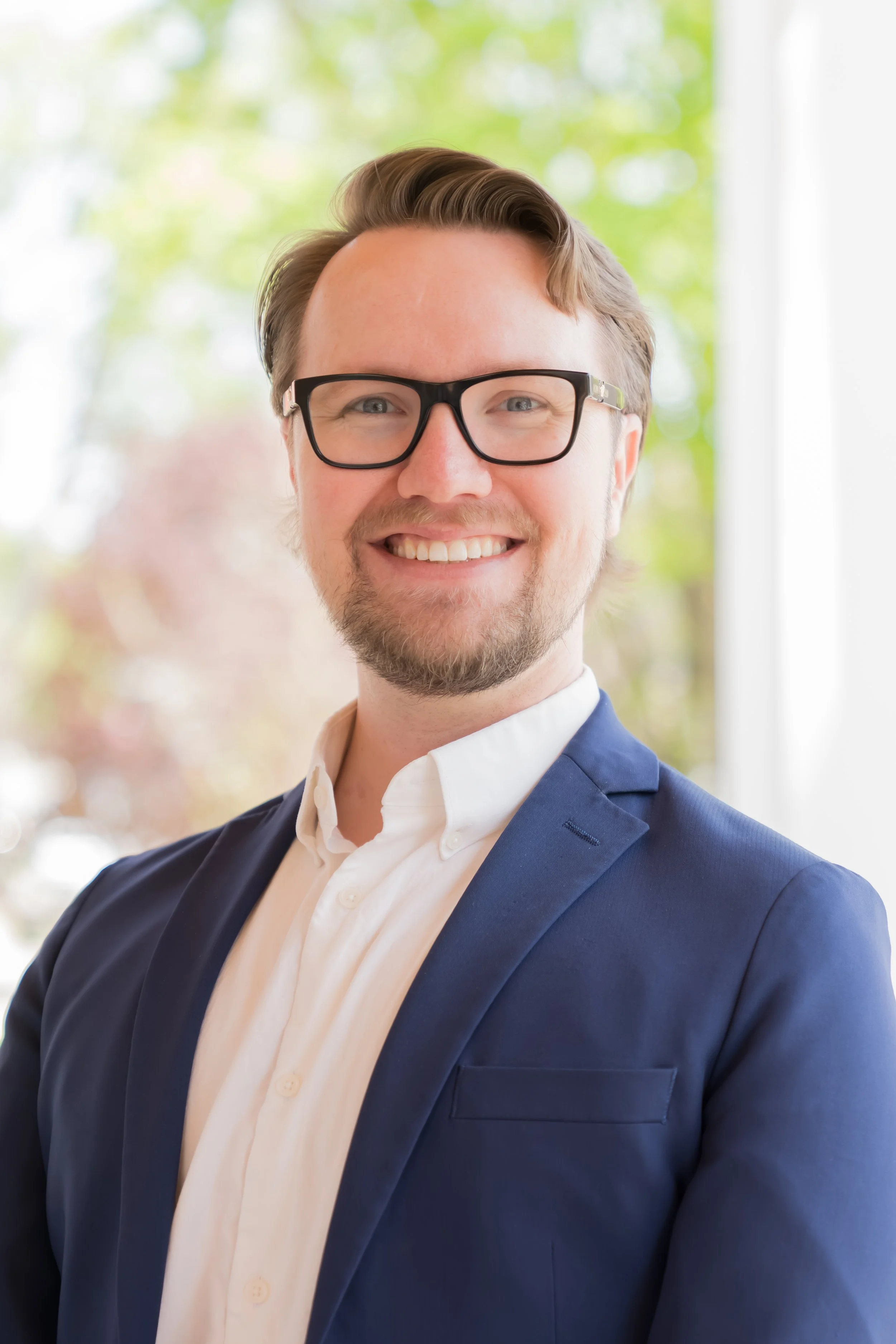 A smiling man with light brown hair, glasses, a beard, dressed in a blue suit jacket, white shirt, and light-colored tie, standing indoors with a background of green trees seen through a window.
