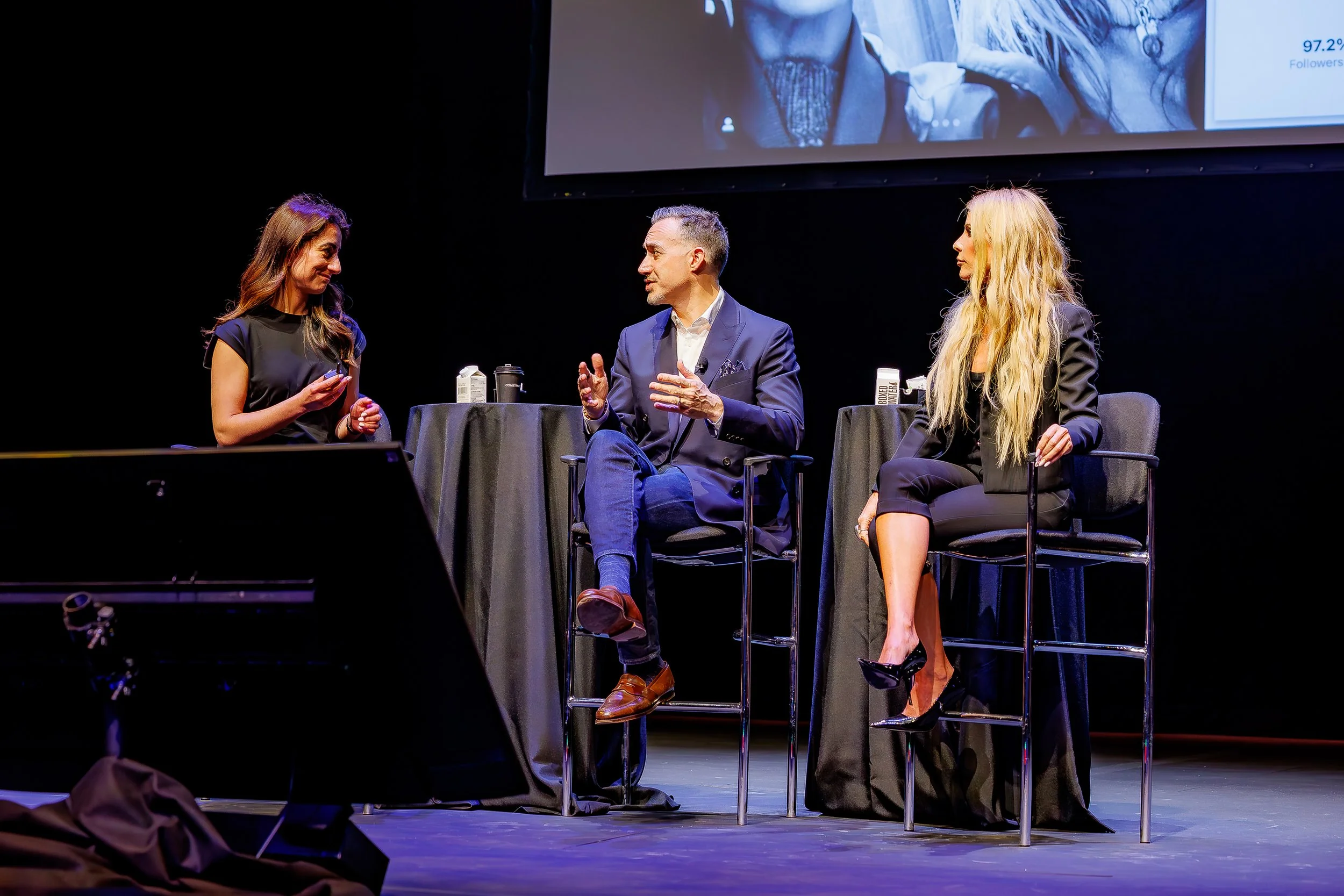 Three people sitting on stage in a discussion, with a large screen overhead. The person in the middle is speaking, wearing a dark suit. The two women on either side are listening, dressed professionally. The stage has black tables with items on them.