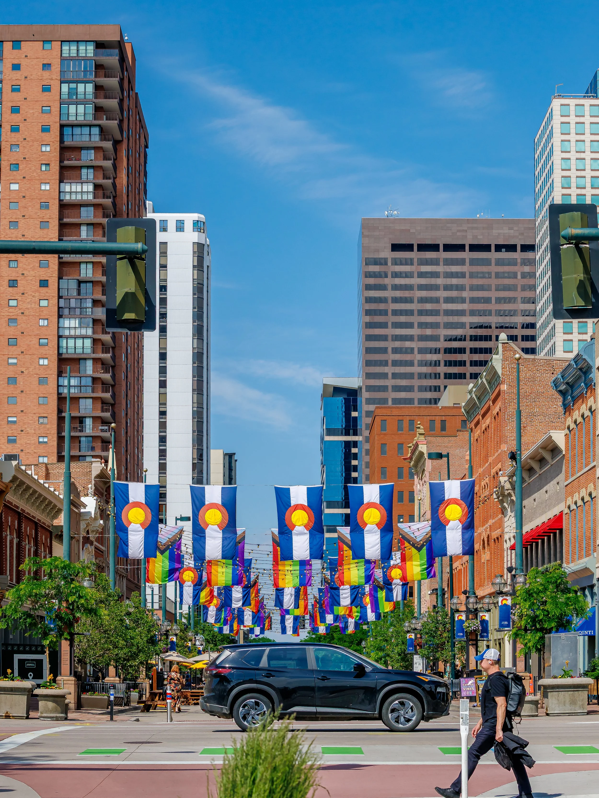 City street decorated with behalf of Colorado flags and rainbow flags, with high-rise buildings in the background, trees on the sidewalks, a black SUV, and a person walking with a backpack.